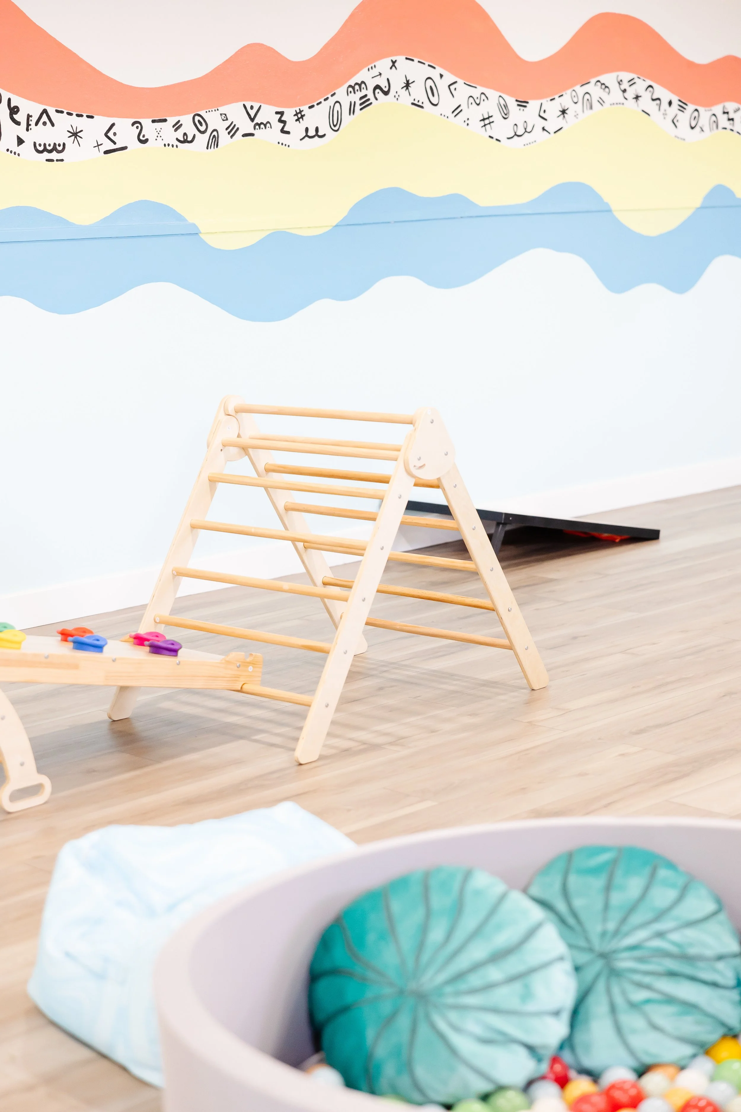 Children's indoor play area with a wooden climbing frame, a ball pit with soft ball cushions, and a colorful abstract mural on the wall.