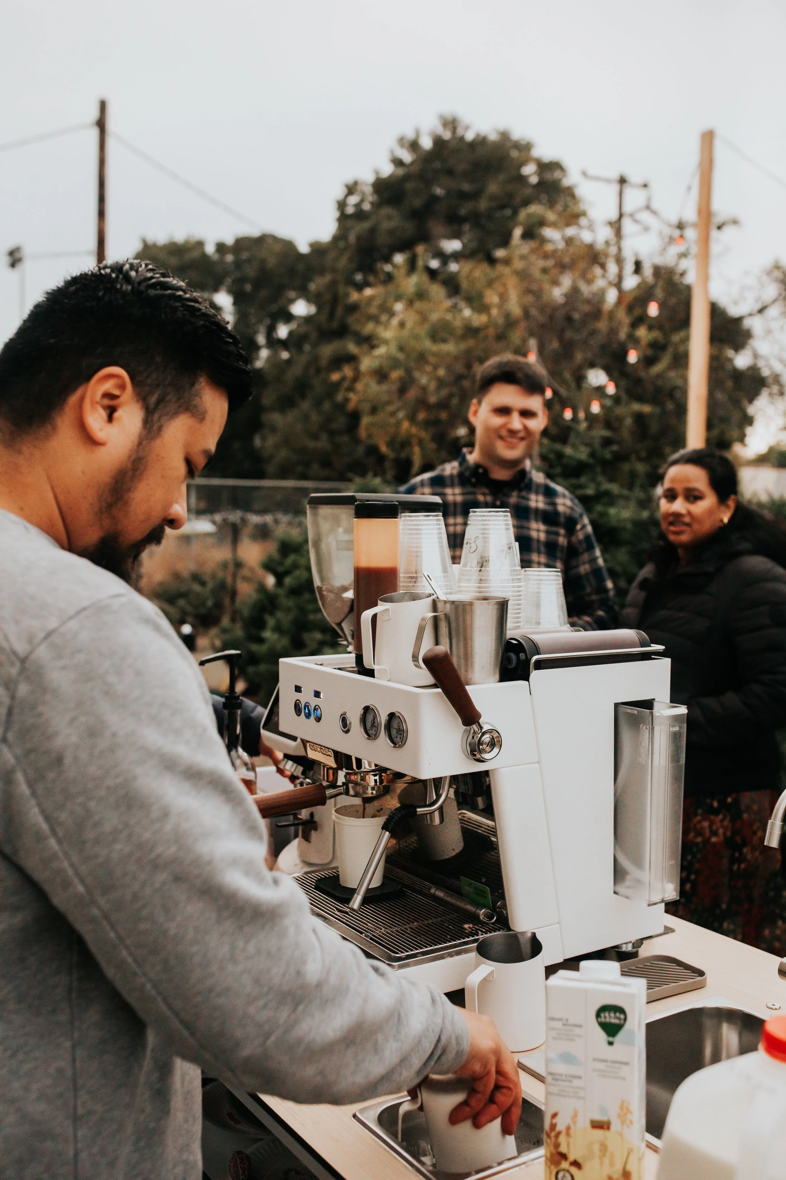 A person making coffee with a commercial espresso machine at an outdoor event, with two people smiling in the background against a natural setting with trees and utility poles.