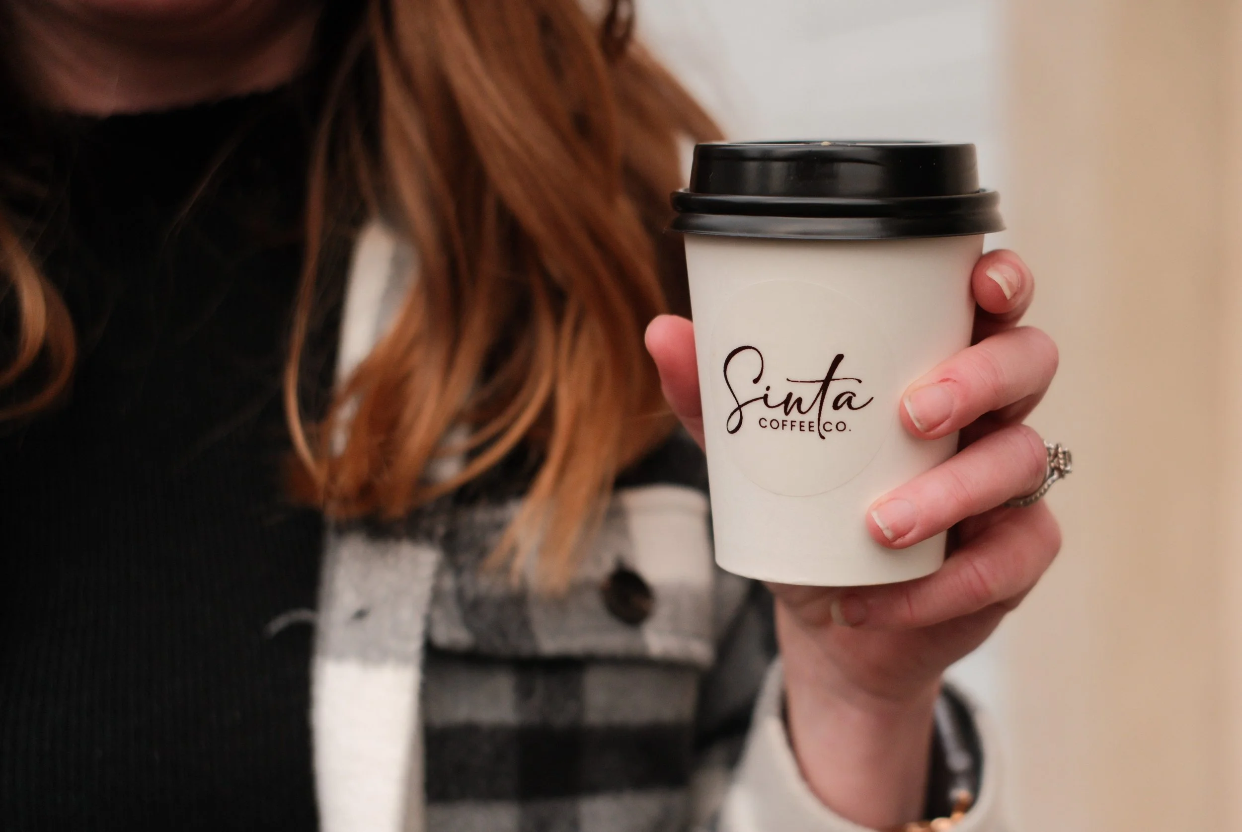 A person with red hair holding a white paper coffee cup with a black lid, branded with 'Sinta Coffee Co.'