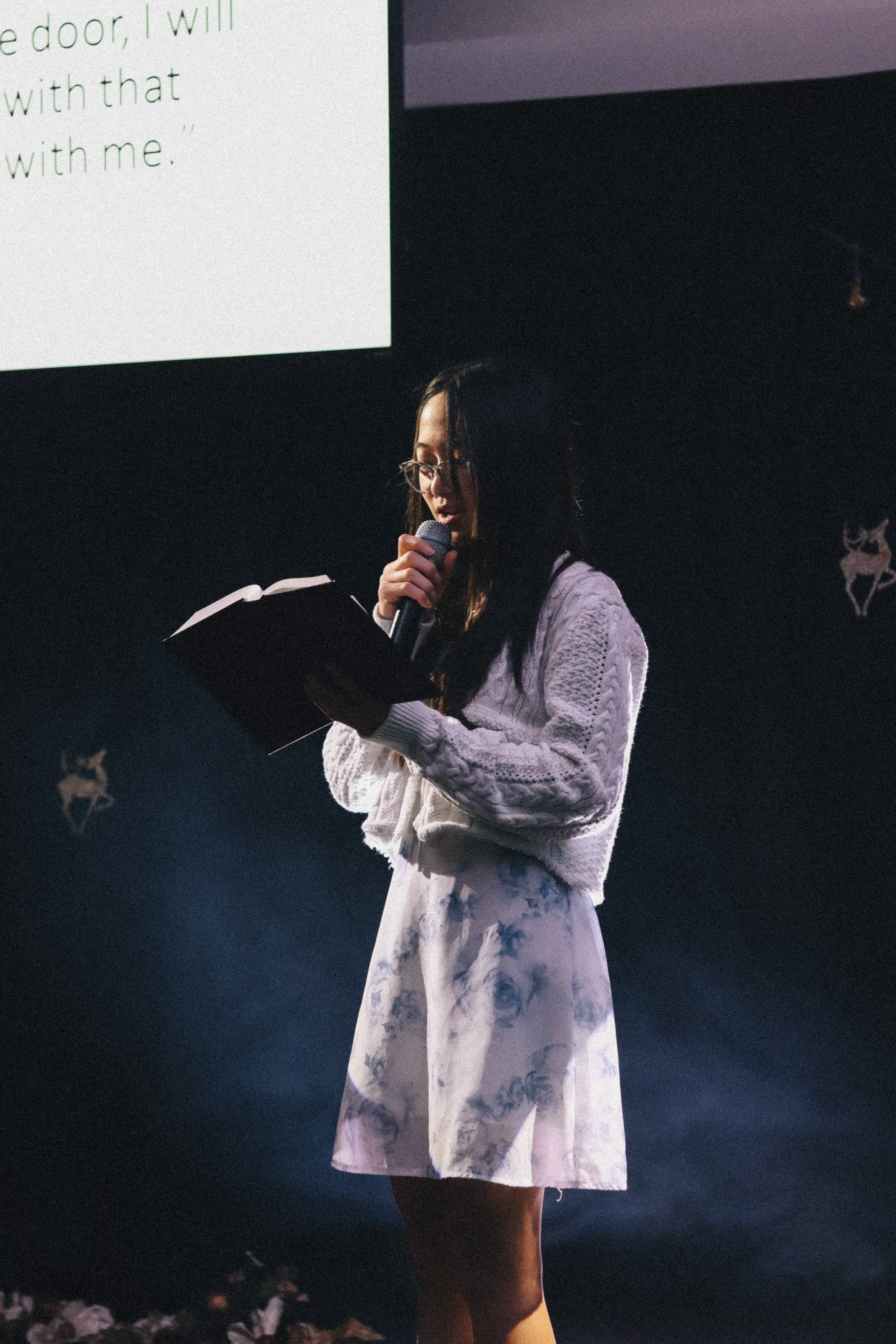 A woman reading from the Bible into a microphone at a dark stage.