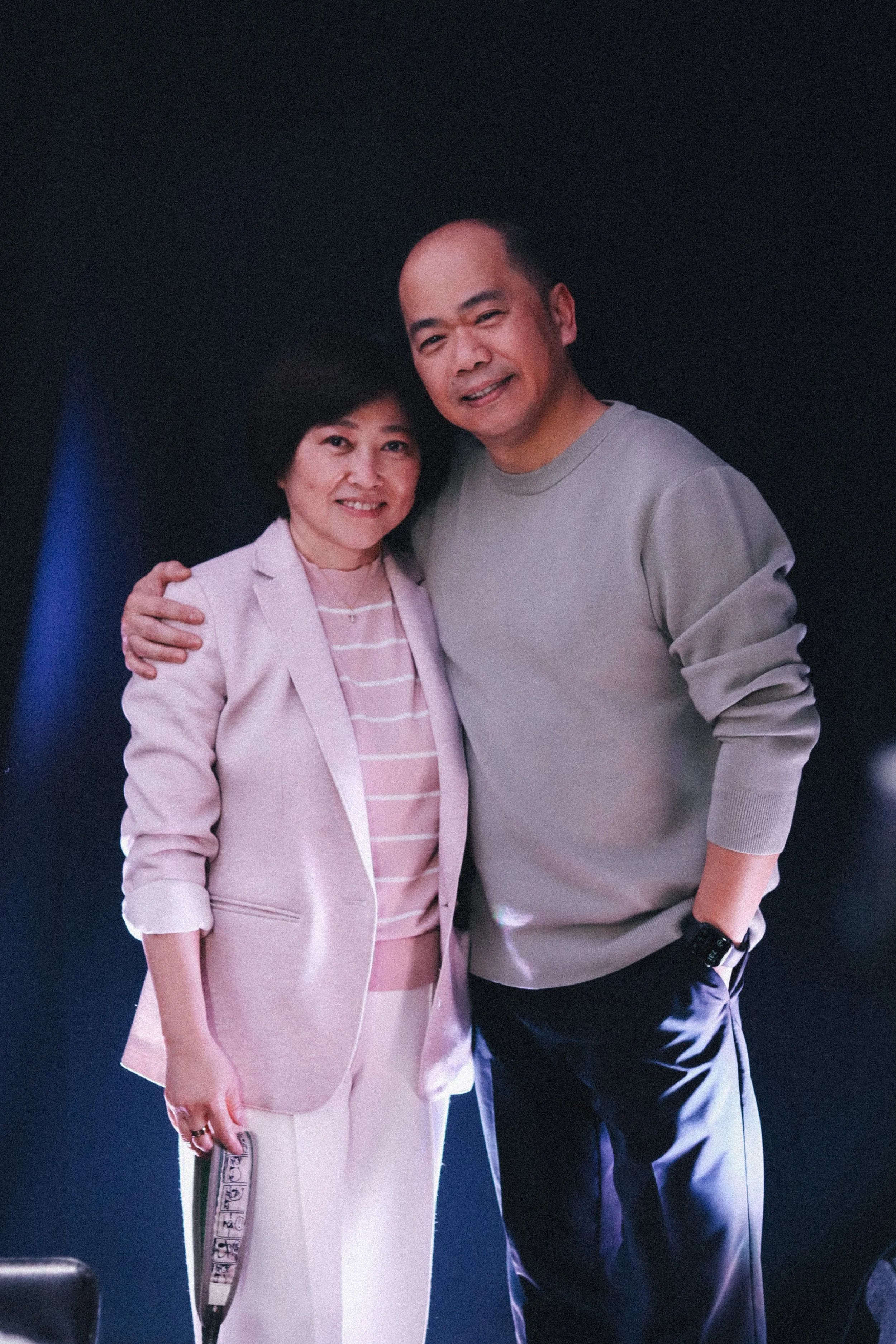 Senior Pastor Devi and his wife Juni posing together, smiling and standing close with their arms around each other against a dark background.