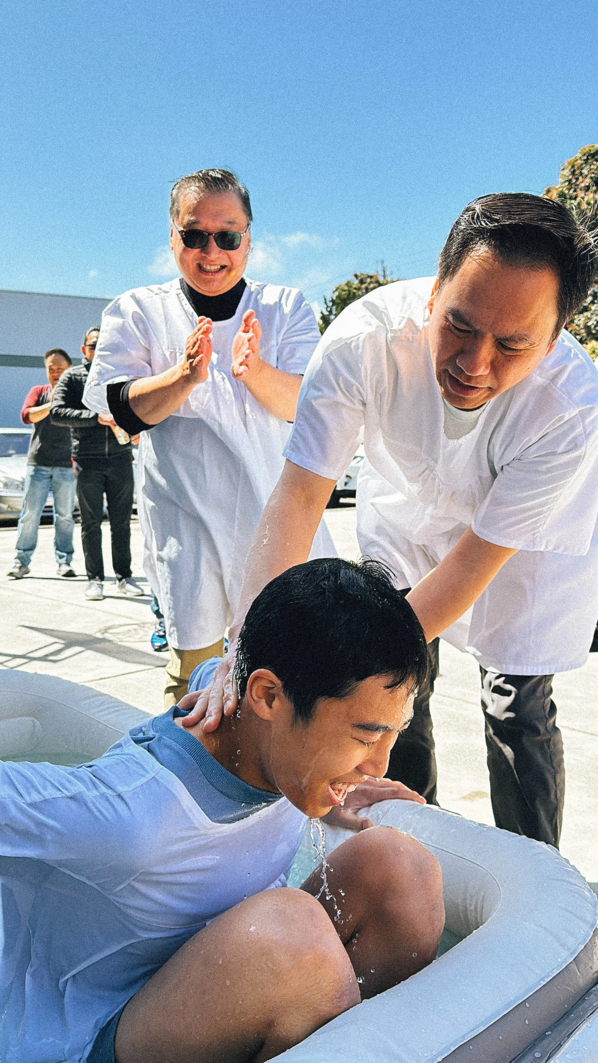 Two Pastors blessing in a baptism ceremony outdoors in a small pool, with a group of people observing and celebrating in the background, under a clear blue sky.