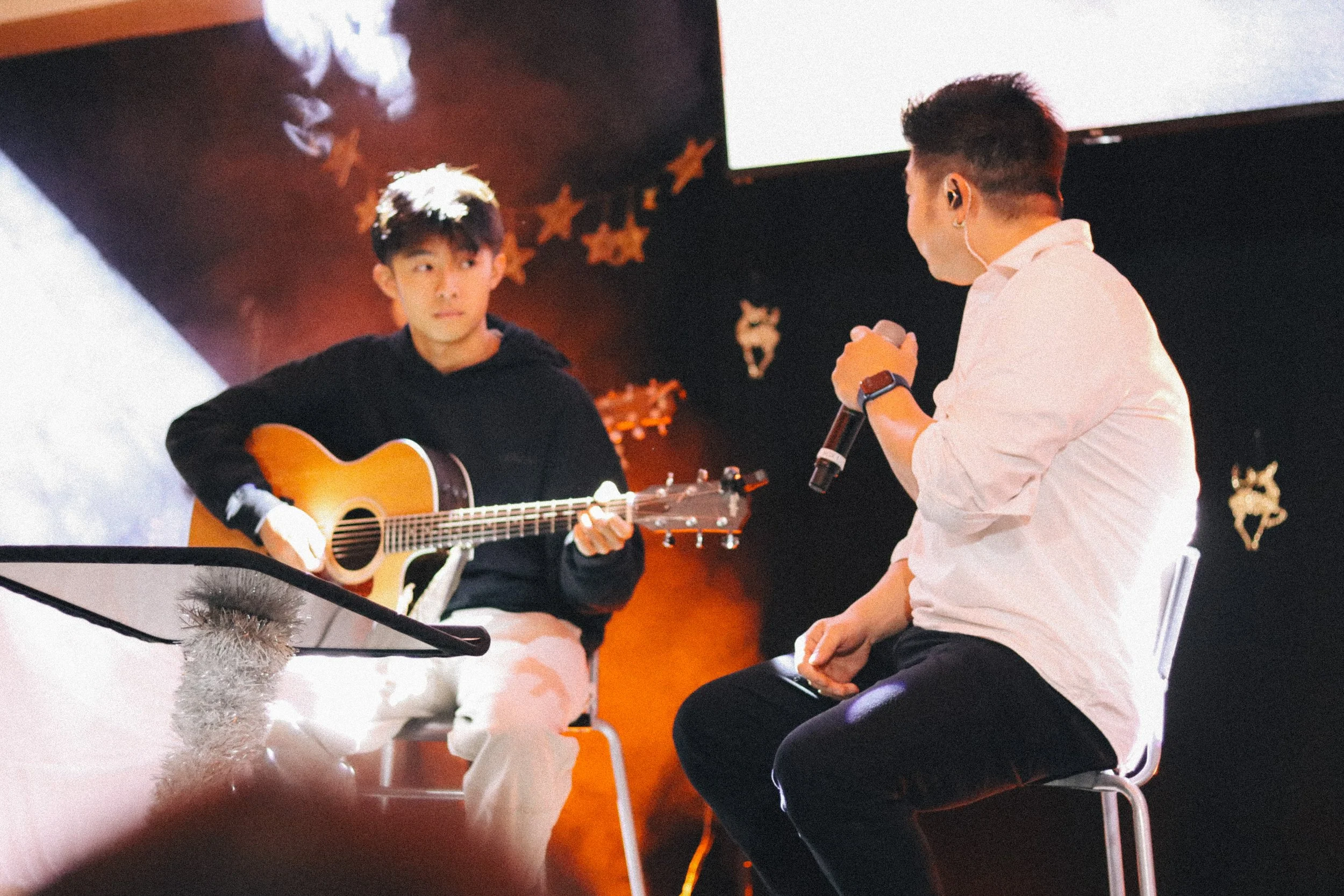 A young man is playing an acoustic guitar on stage, while another young man, sitting on a stool, is holding a microphone singing during Christmas service. The background features holiday-themed decorations, including stars and reindeer.