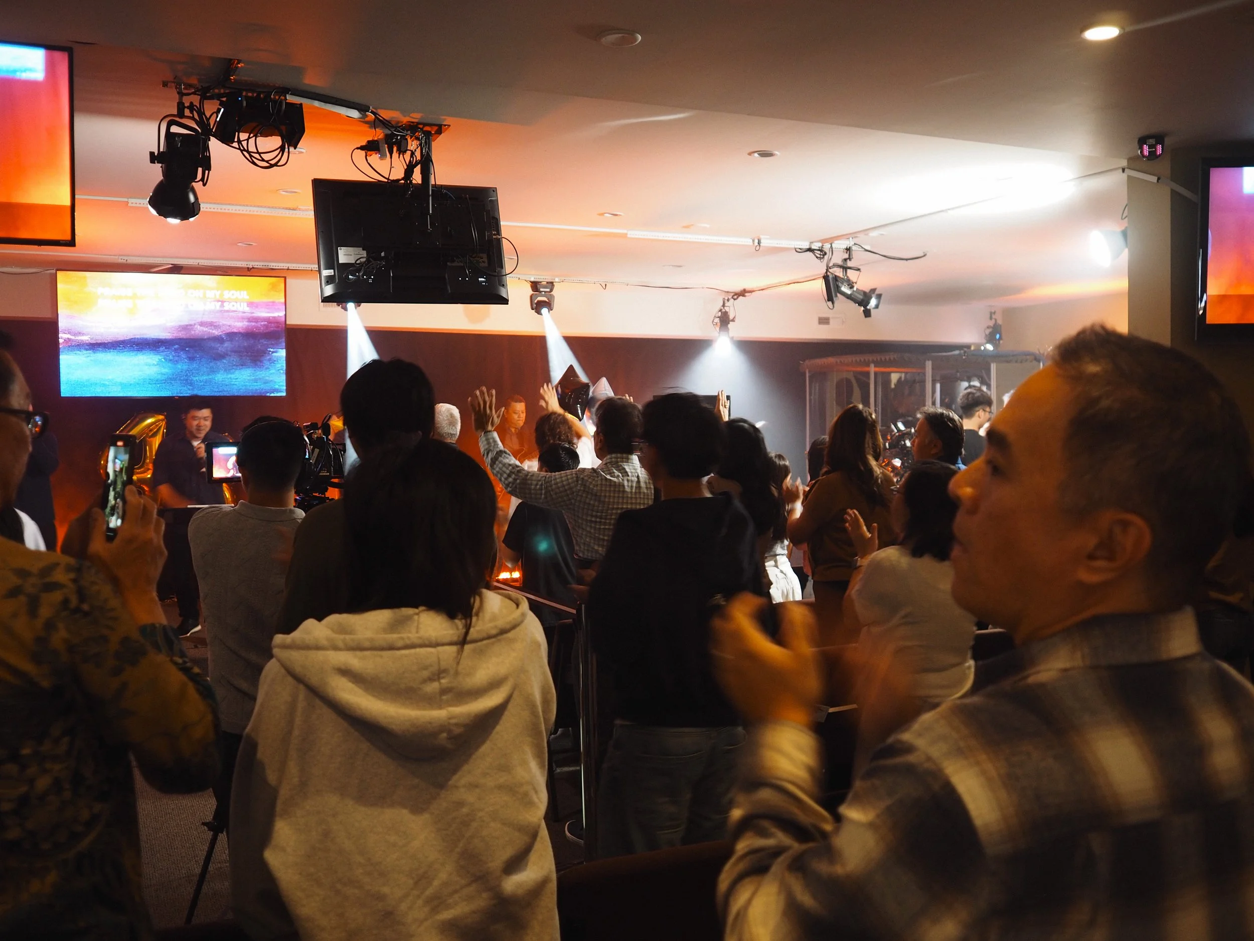 People are gathered indoors, attending a worship event with a stage, lighting, and large screens displaying colorful backgrounds.