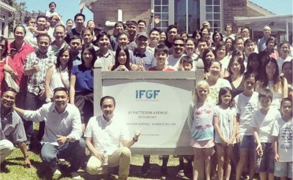 Group of diverse people, including children and adults, gathered outdoors for a photo with a sign that reads 'IFGF Auckland Church' in front of a modern building.