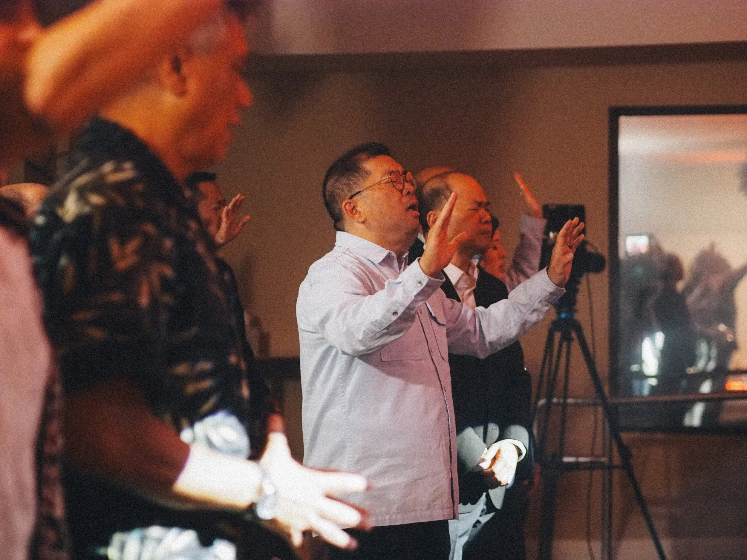 Group of people at a Sunday service with their eyes closed and hands raised in prayer or worship in a dimly lit room.