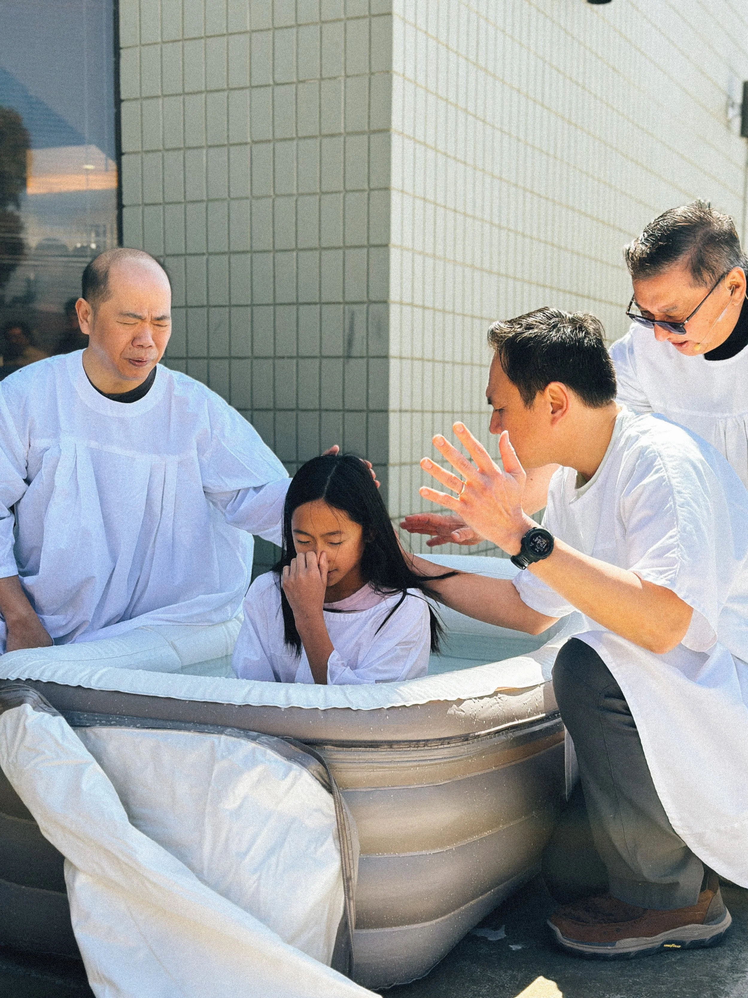 A girl being baptized in an outdoor inflatable pool by several Pastors dressed in white robes, with the church wall in the background.