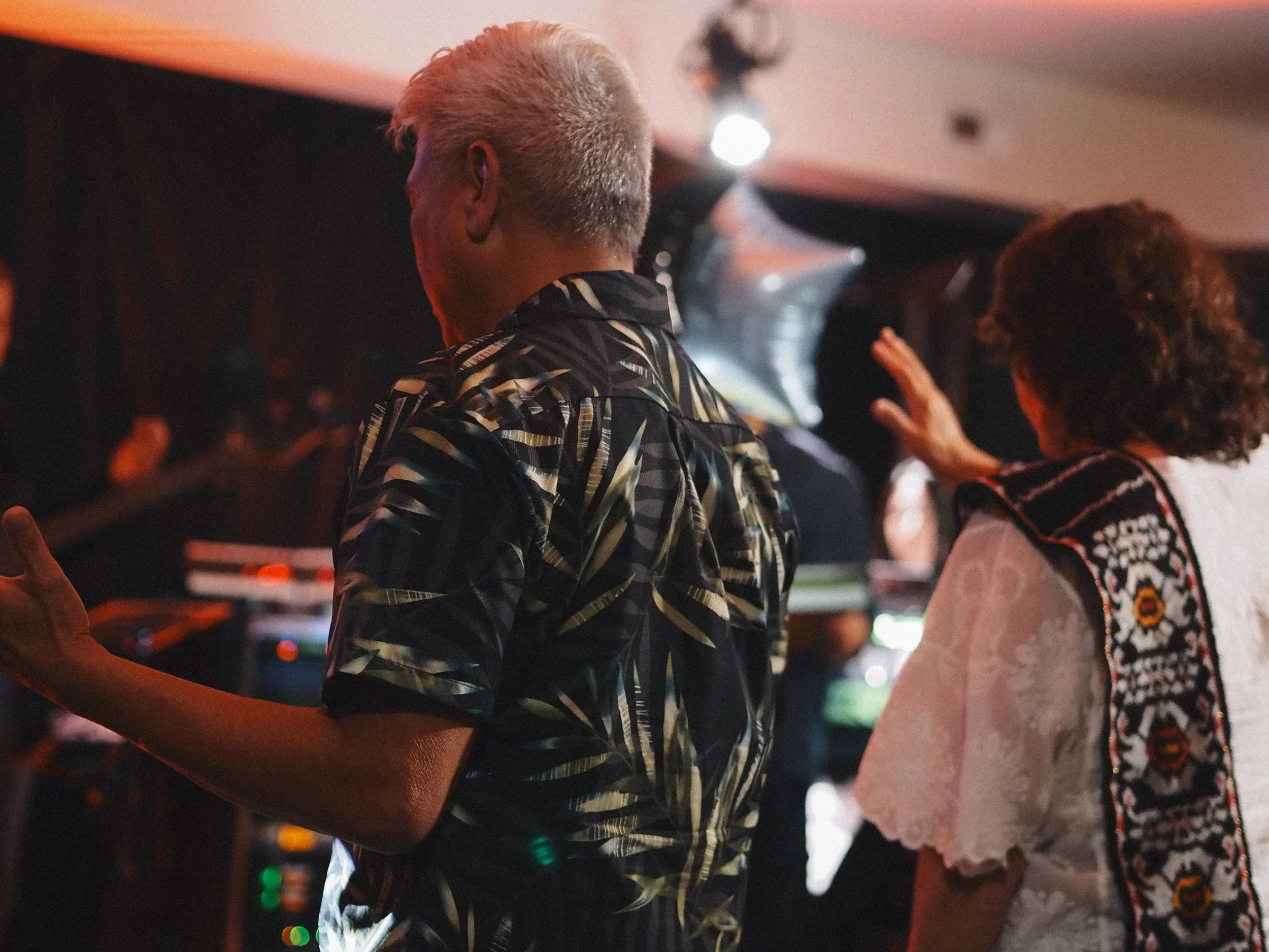 Two people are seen from behind at a Sunday Service, one with short, gray hair wearing a patterned shirt, and the other with curly brown hair wearing a white garment with embroidered details, gesturing with their hand.