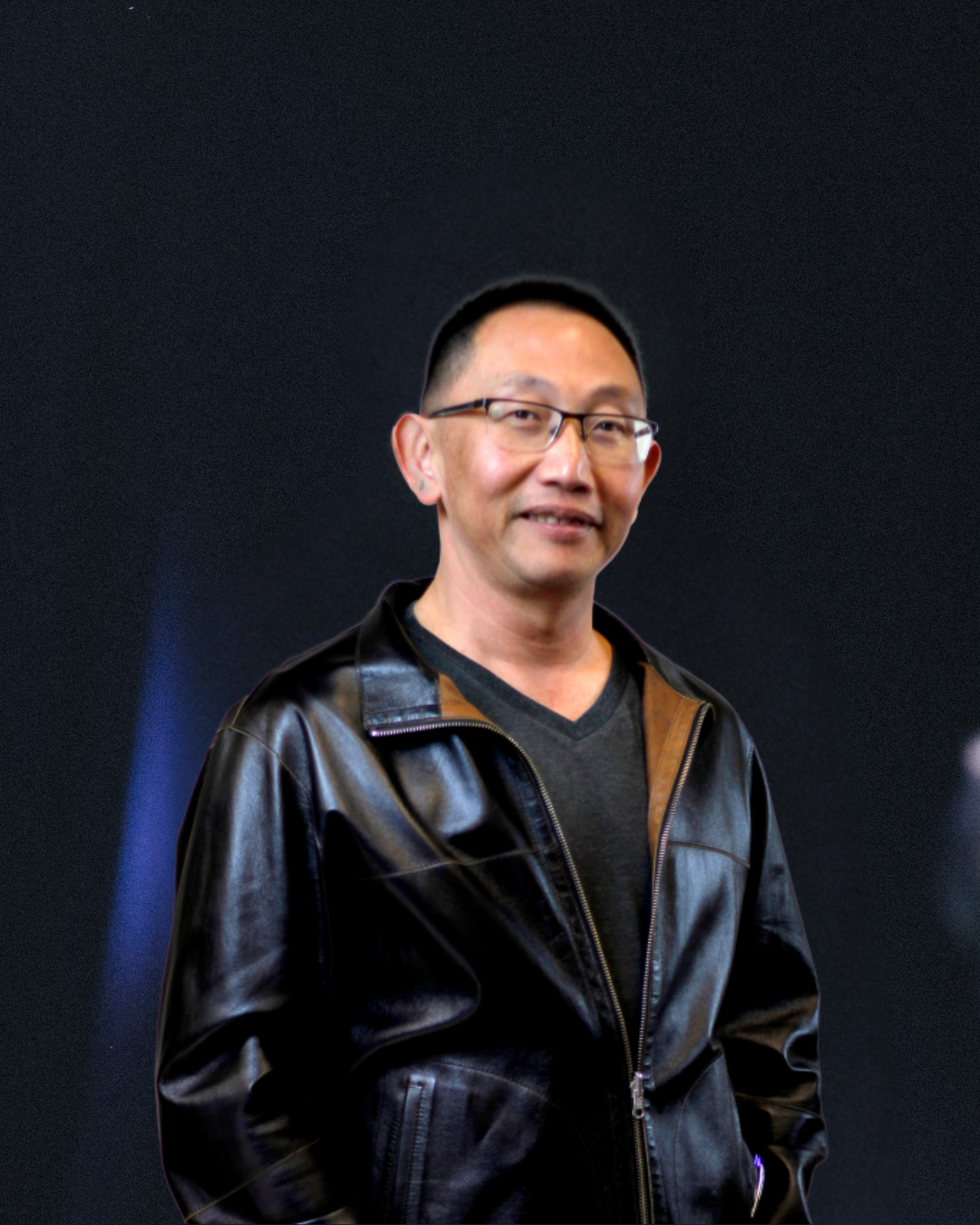 Associate Pastor Deky wearing a black leather jacket stands against a dark background.