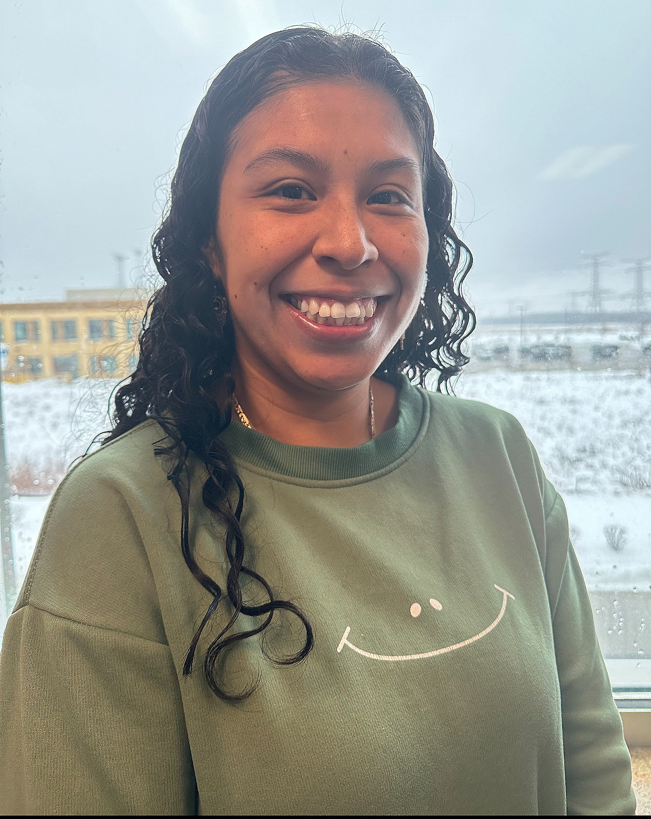 A smiling woman with curly black hair wearing a green sweatshirt with a white smiling face graphic, standing in front of a window with a snowy landscape outside.