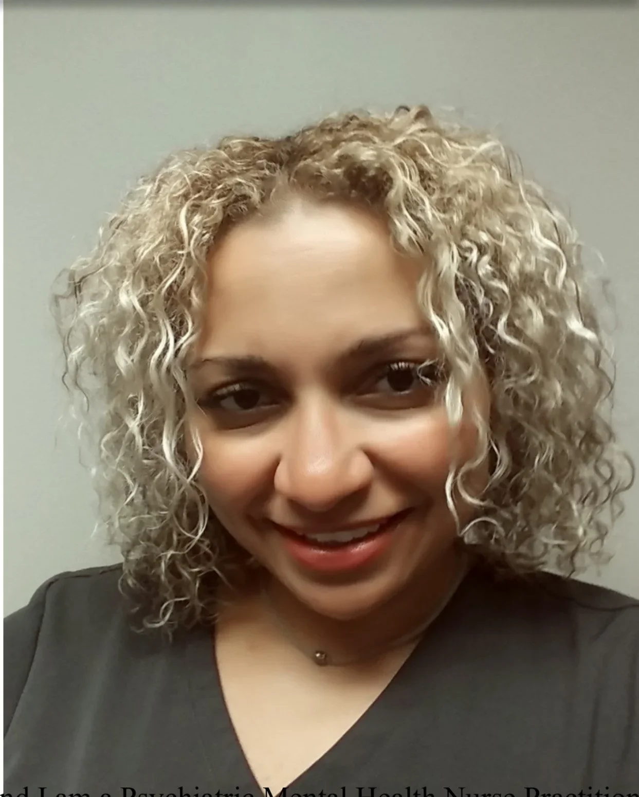 A woman with curly blonde hair smiling at the camera, wearing a dark top and a necklace.