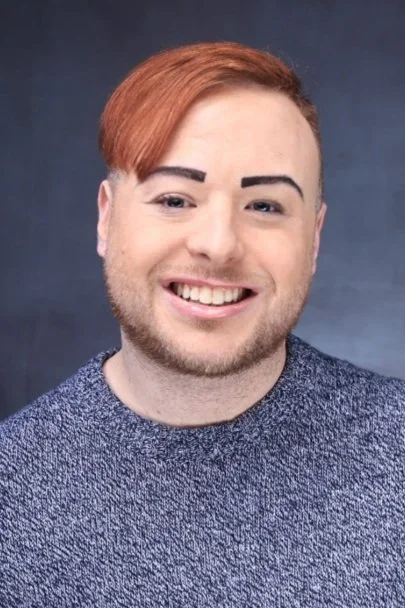 A smiling young man with red hair, a beard, and a mustache, wearing a textured gray shirt, against a dark background.