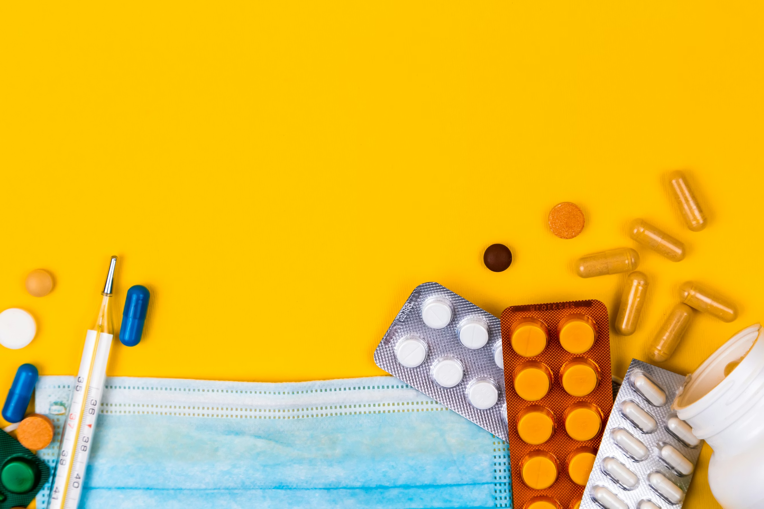 Medical items including a face mask, various blister packs of pills, capsules, and tablets, and a thermometer scattered on a yellow background.