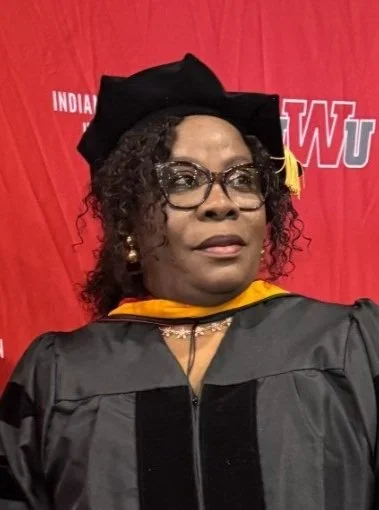 Woman wearing academic regalia and a cap, standing in front of a red backdrop with university logos.