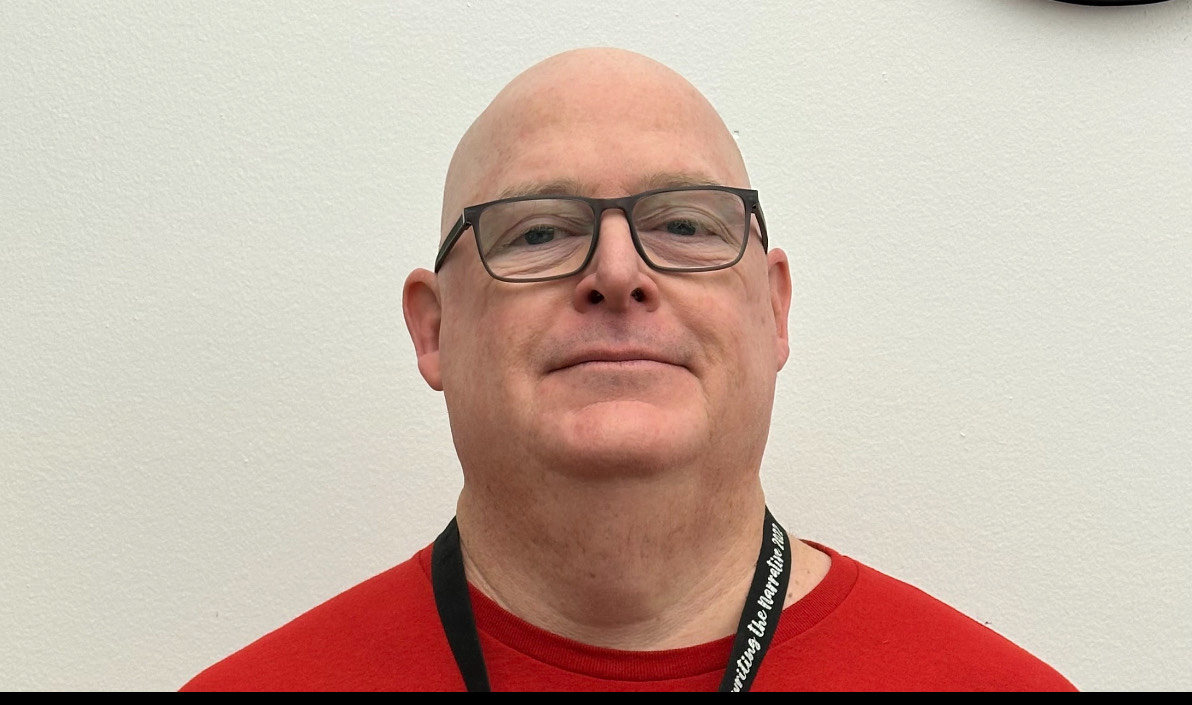 A bald man wearing black glasses and a red shirt standing against a plain light-colored wall.