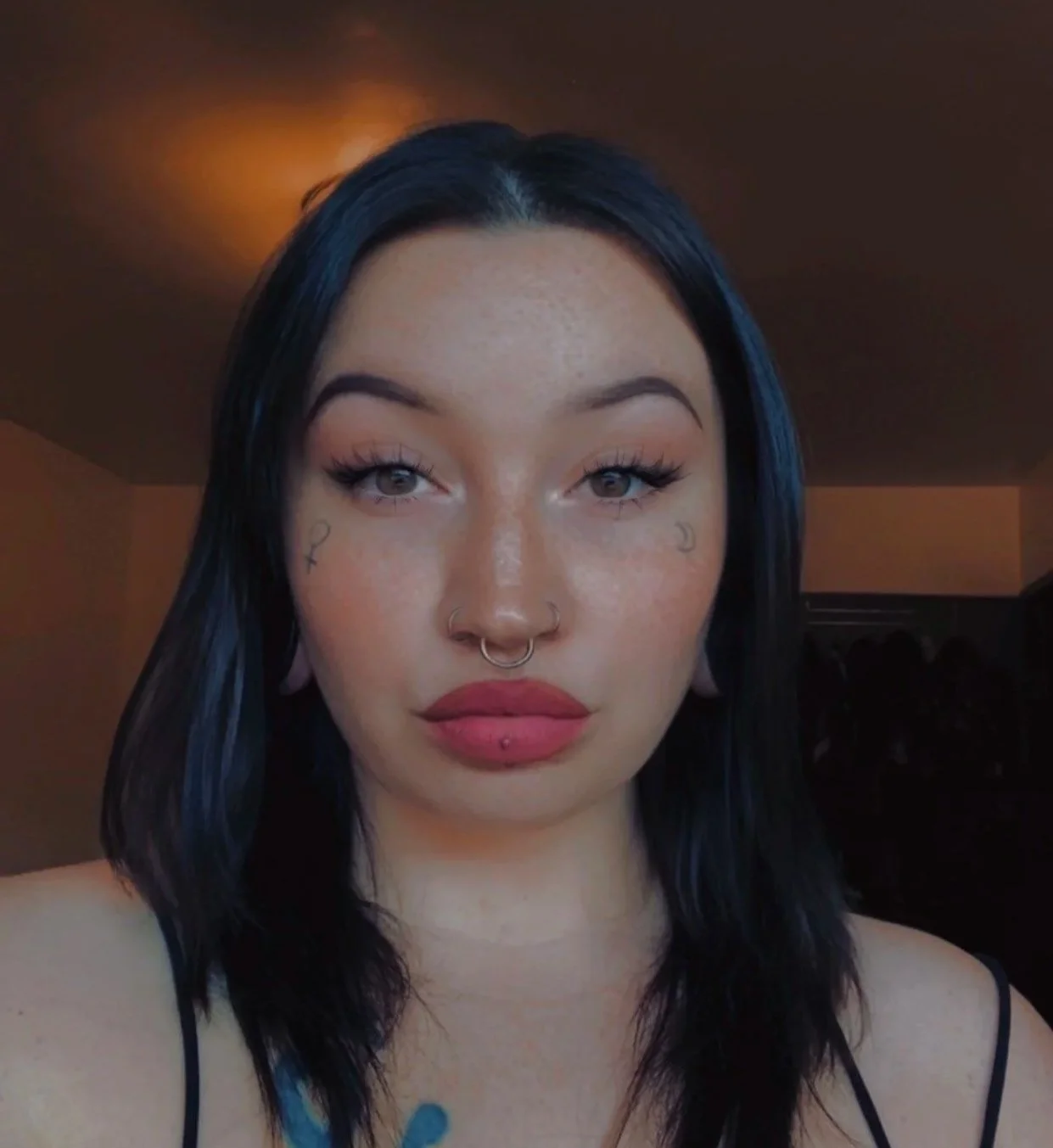 A young woman with black hair, light brown eyes, and multiple facial piercings, including septum, labret, and nostril piercings, taking a selfie indoors with warm lighting.