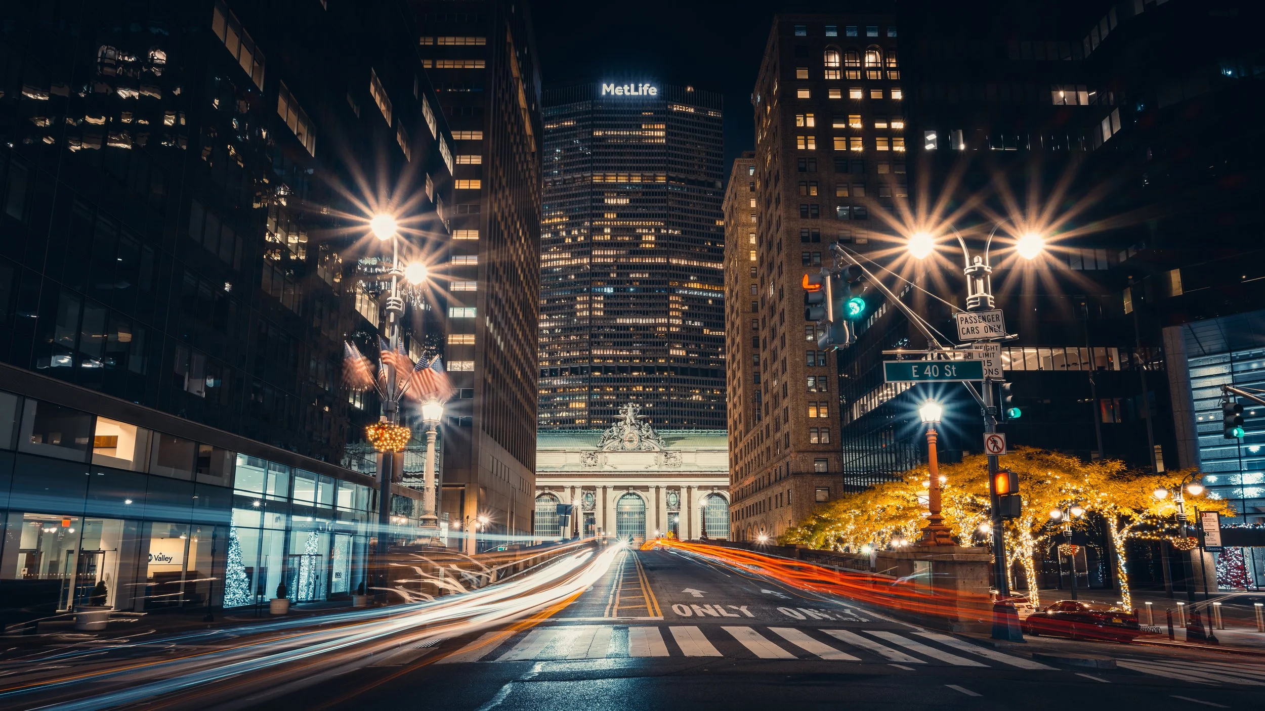 Nighttime city street scene in New York City with tall illuminated skyscrapers, streetlights, and blurred moving cars, including the MetLife building in the background.