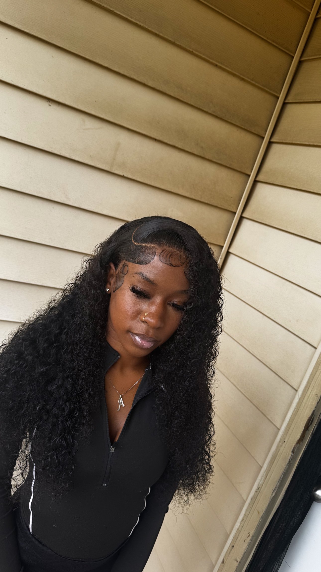 A woman with long curly black hair, wearing a black zip-up jacket with white accents, a gold nose ring, and a necklace with the letter 'A', looking down with a neutral expression against a beige wooden wall background.