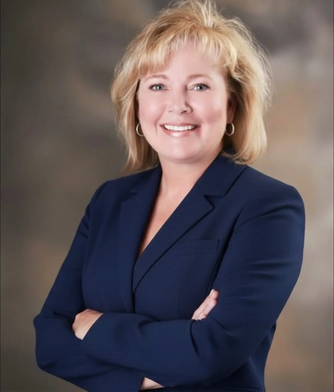 A professional woman with shoulder-length blond hair and blue eyes, wearing a navy blazer and small hoop earrings, smiling with arms crossed against a blurred gray background.