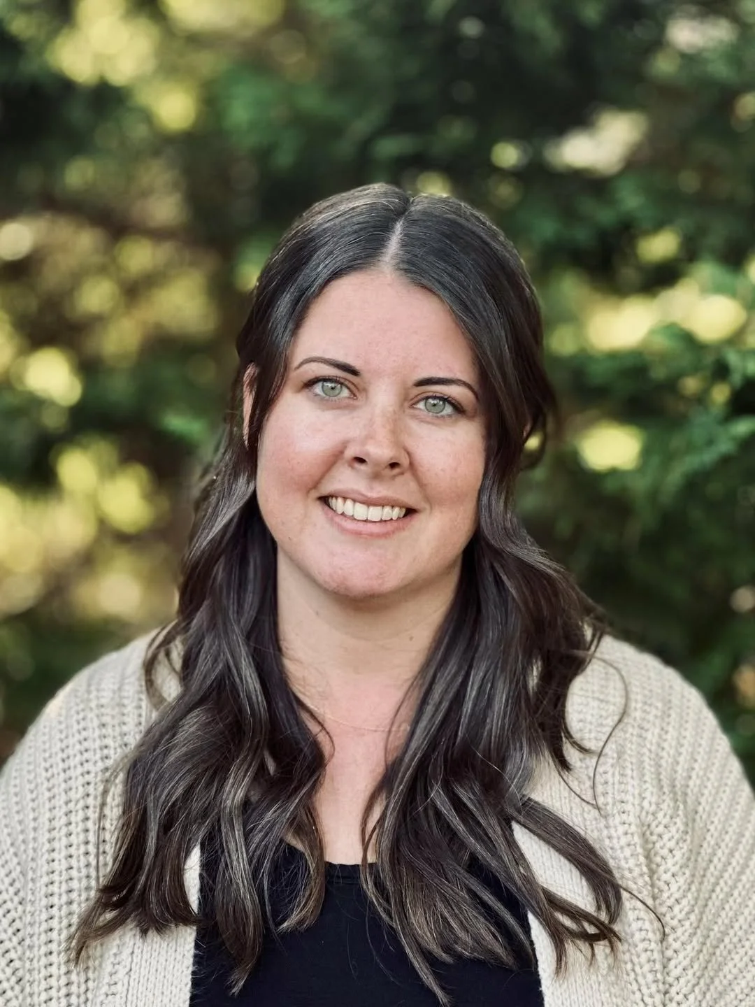 Portrait of a woman with long dark hair and blue eyes, smiling, outdoors with green blurred foliage background.