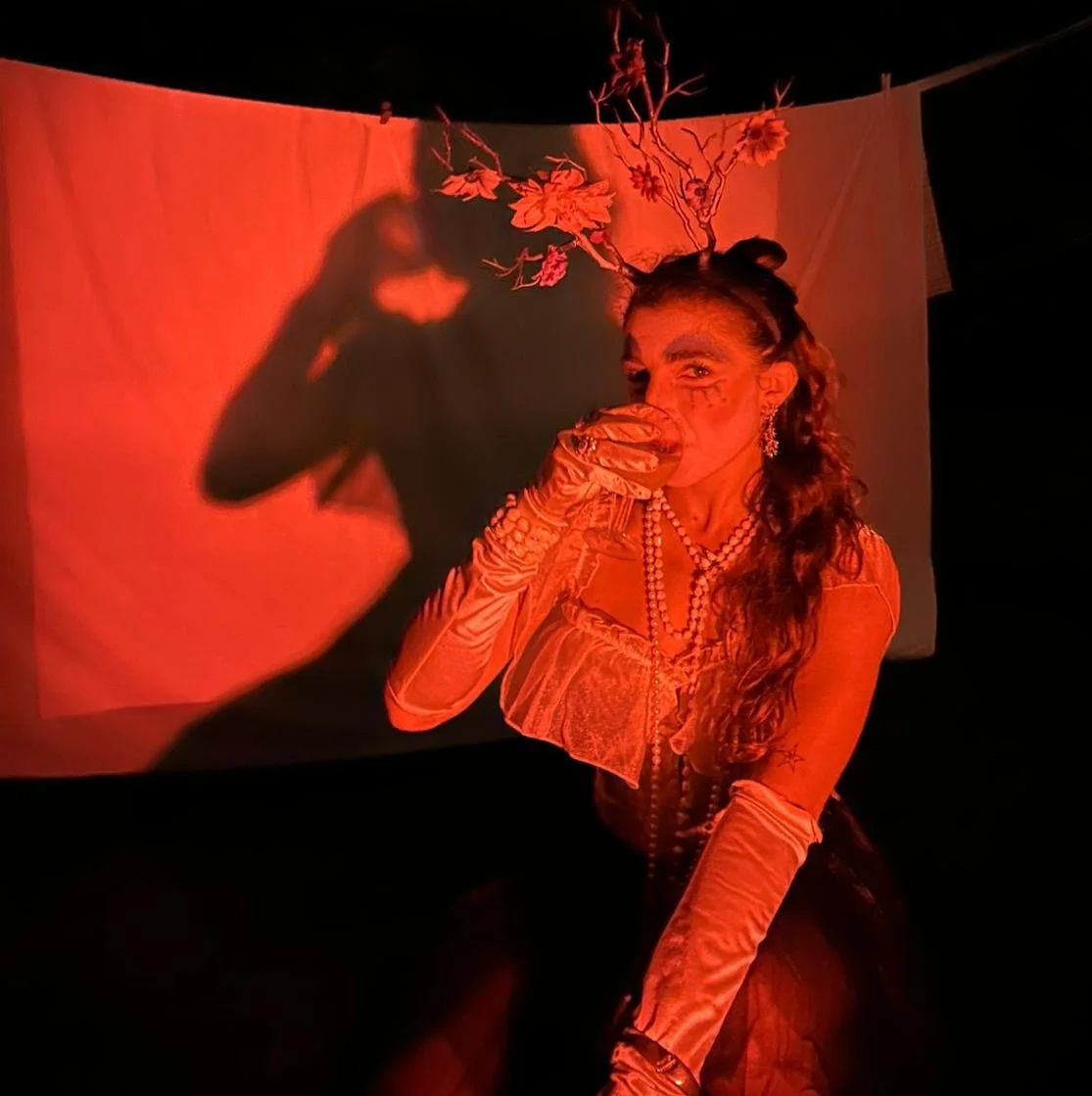 Une femme déguisée en créature forestière avec des bois et des fleurs sur la tête, portant des bijoux et une robe ancienne, sous une lumière rouge, avec une ombre sur un fond blanc.