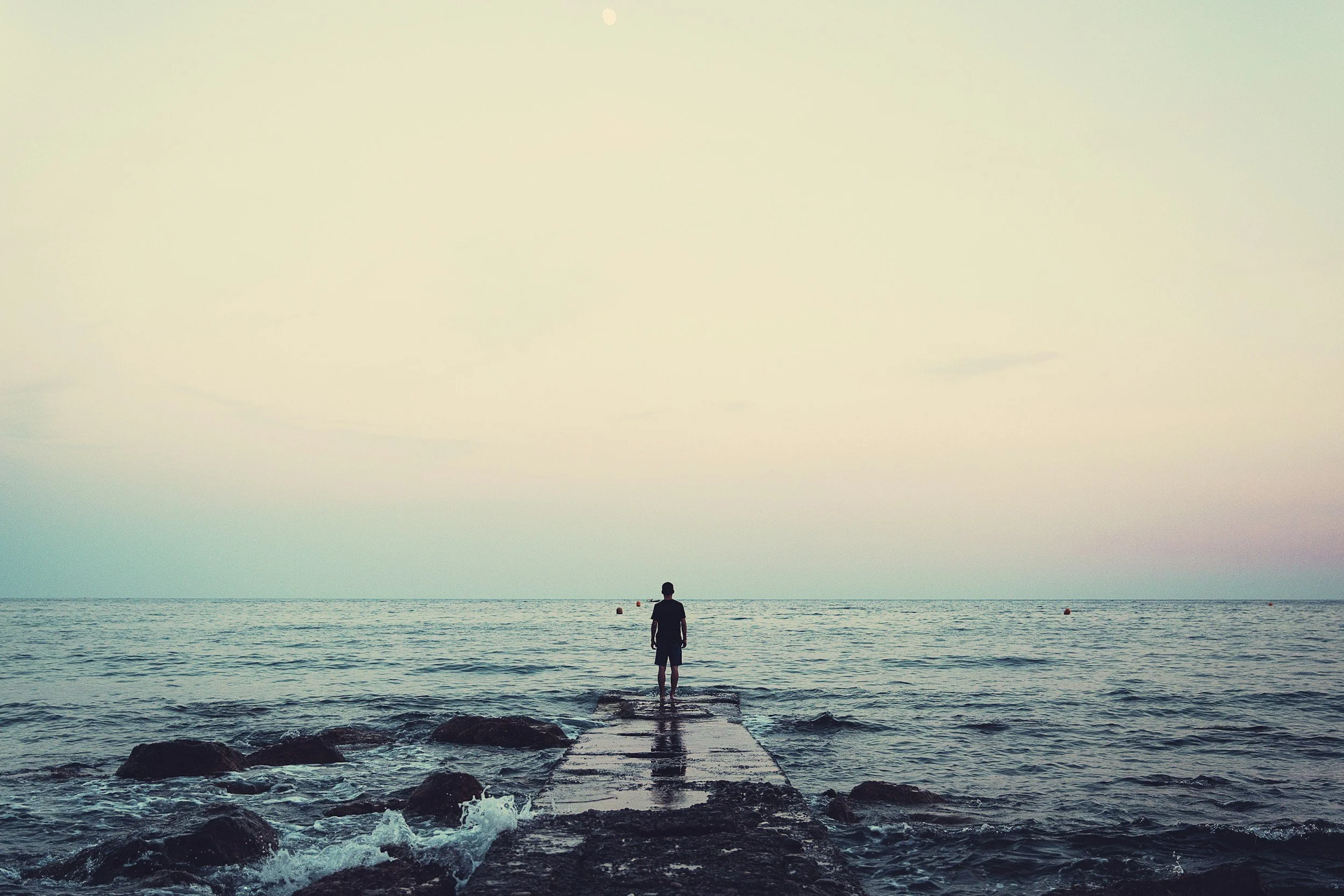A person standing on a pier overlooking the ocean during sunset or twilight, with a calm sea and a pastel-colored sky.
