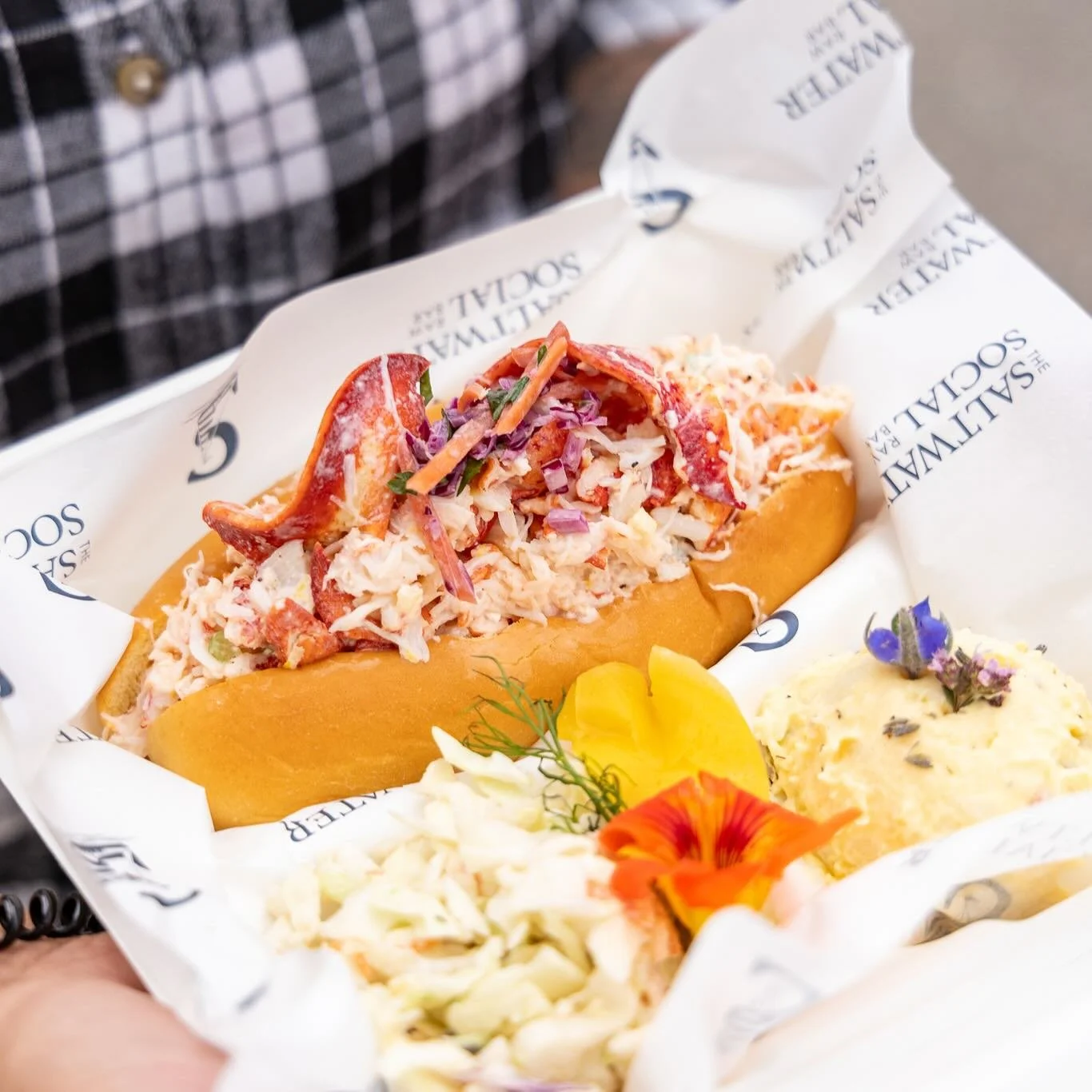 🦞✨ Meet your new coastal craving &mdash; our signature Lobster Roll is piled high with fresh Atlantic lobster, tossed in a light, creamy dressing, and topped with a crisp house-made slaw. Served with vibrant sides and a little edible flower flair 🌸