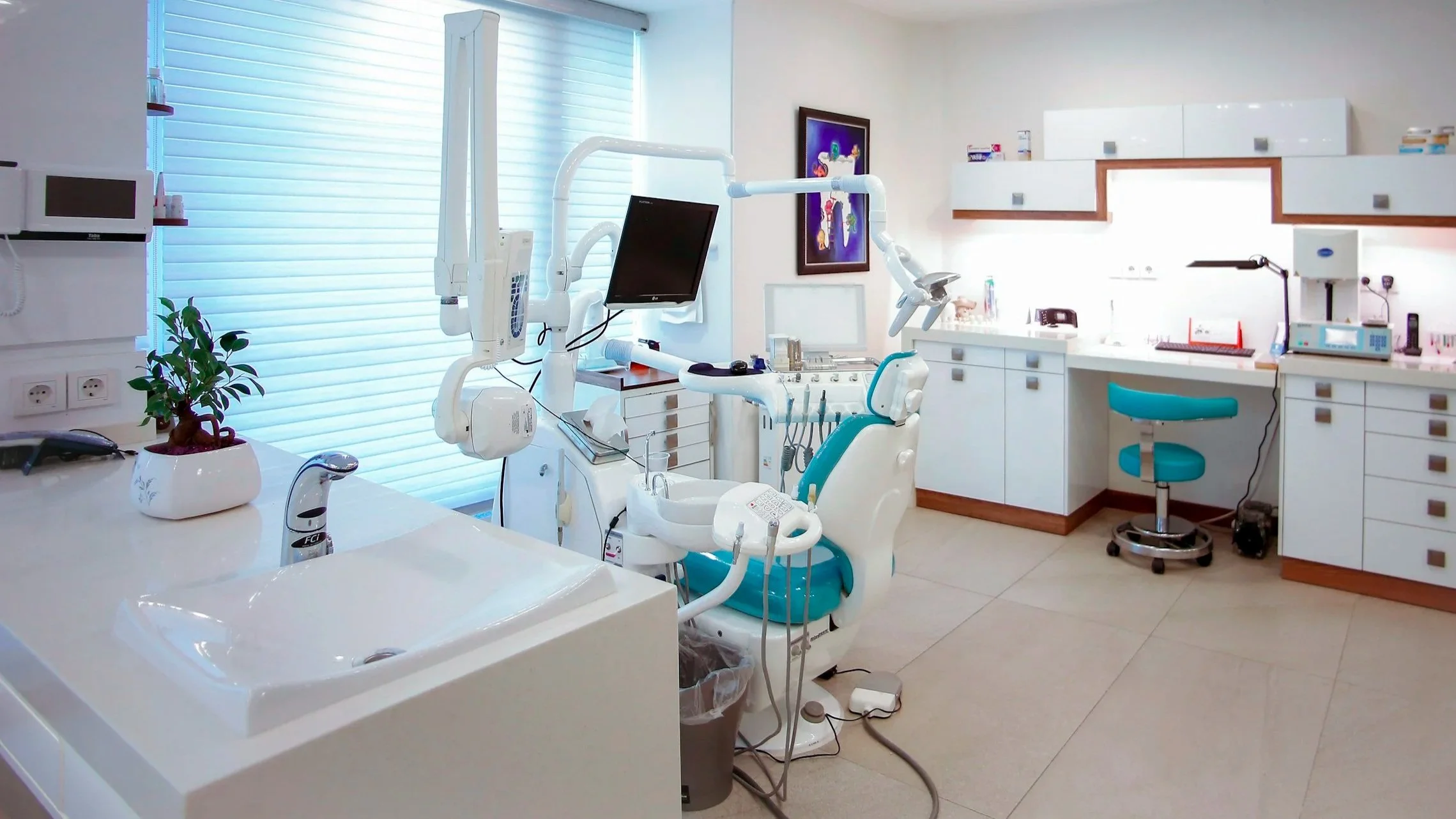 A modern dental clinic with a dental chair, computer monitor, dental tools, a sink, and various cabinets and equipment. The space is clean and well-lit with natural light coming through window blinds.
