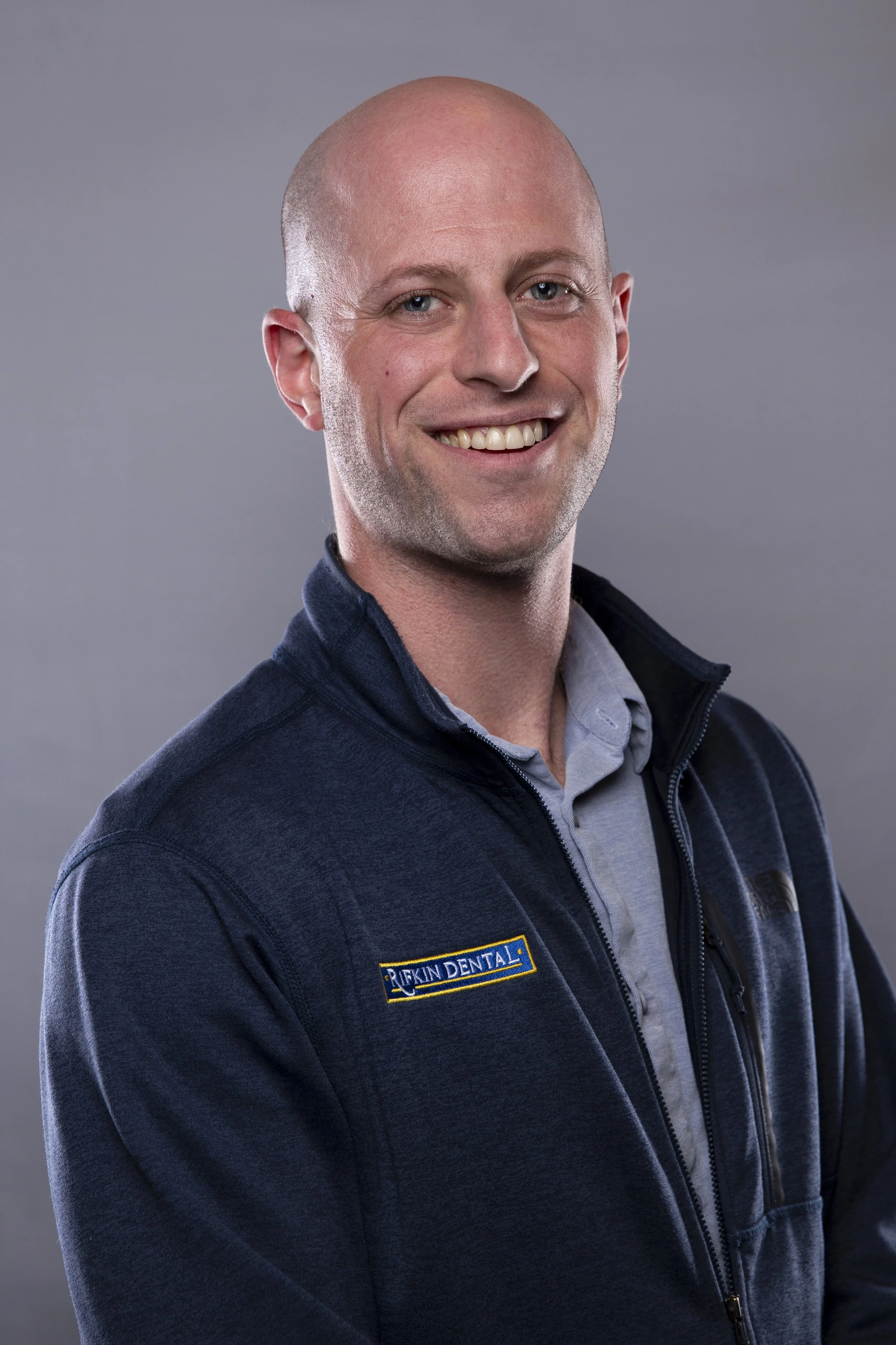 Portrait of a smiling man with a shaved head wearing a blue zip-up jacket with a patch that reads 'Rifkin Dental' over a light gray shirt.