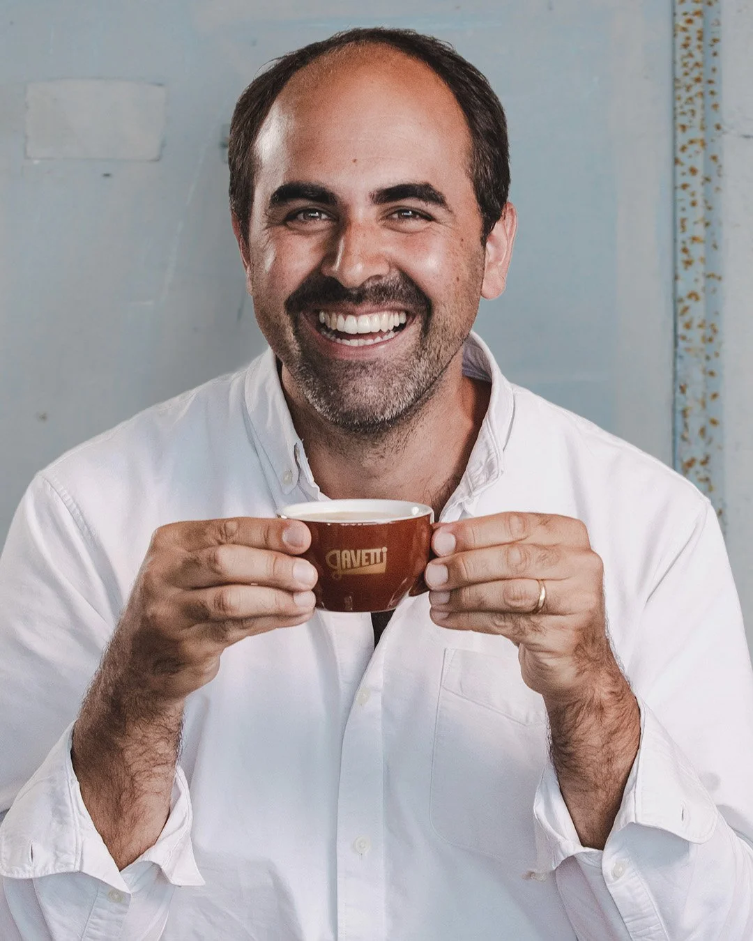 A smiling man holding a small brown coffee cup with both hands, wearing a white button-down shirt, standing against a plain background.