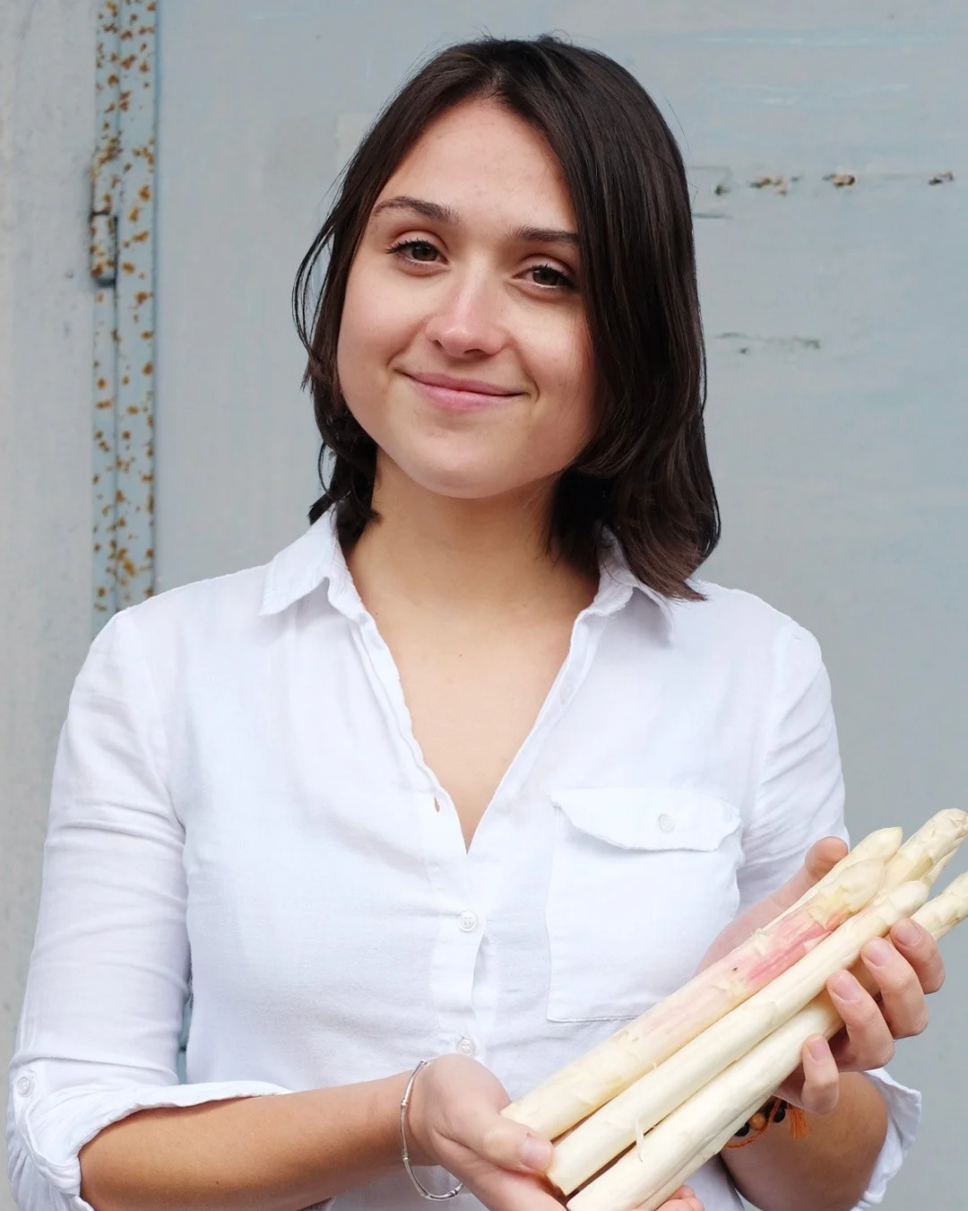 A young woman with shoulder-length dark hair, wearing a white button-up shirt, smiling and holding several white asparagus stalks in her hands.