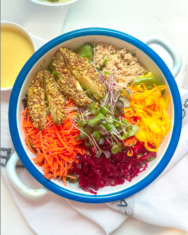 Colorful vegetable salad with shredded carrots, beets, yellow squash, microgreens, quinoa, sliced cucumbers, and stuffed grape leaves topped with diced pistachios, served in a white bowl with a blue rim.
