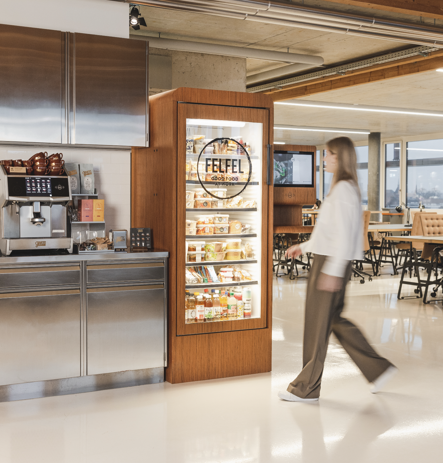 A woman walking past a refrigerated display case in a modern cafe or food court. The display case has a sign that reads 'FELFEL-GOOD FOOD AT WORK' and contains various packaged food items and drinks. The cafe has stainless steel appliances and a seating area with tables and chairs.