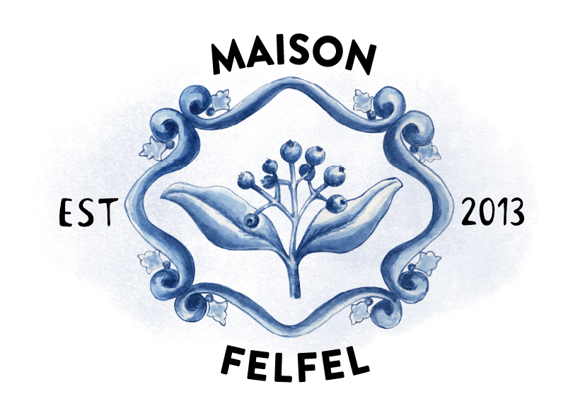 A logo featuring a stylized branch with leaves and berries enclosed by an ornate swirling border, on a dark blue background, with text elements 'EST' on the left, '2013' on the right, and a partial black text below.