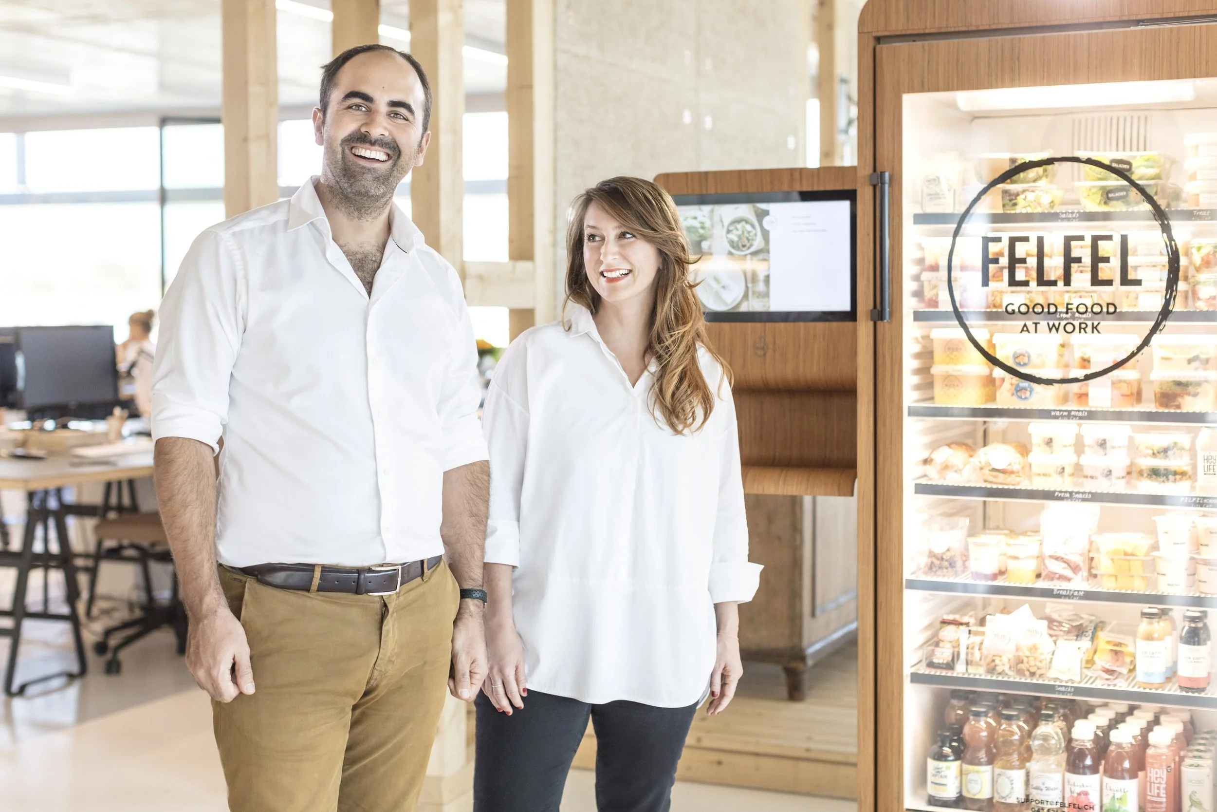 A man and woman standing together inside a modern office or retail space, smiling near a refrigerator filled with various healthy foods, with a sign on the fridge reading 'FELFEL GOOD FOOD AT WORK'.