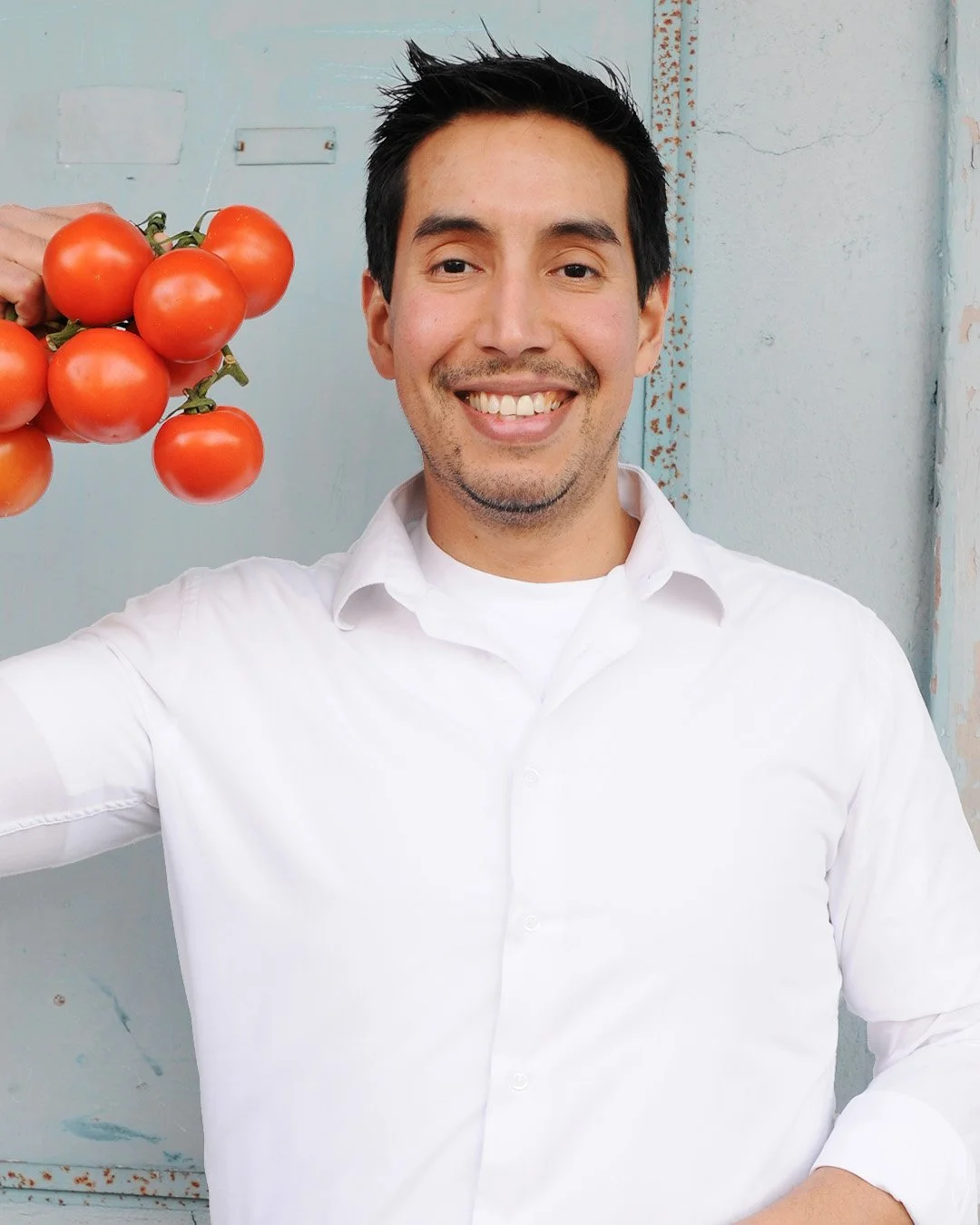 Smiling man in white shirt holding a bunch of red tomatoes, standing against a light blue background.