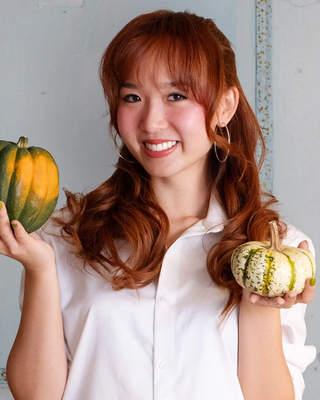 A woman with long red hair, wearing a white shirt and hoop earrings, smiling and holding two small pumpkins, one in each hand, in front of a light-colored background.