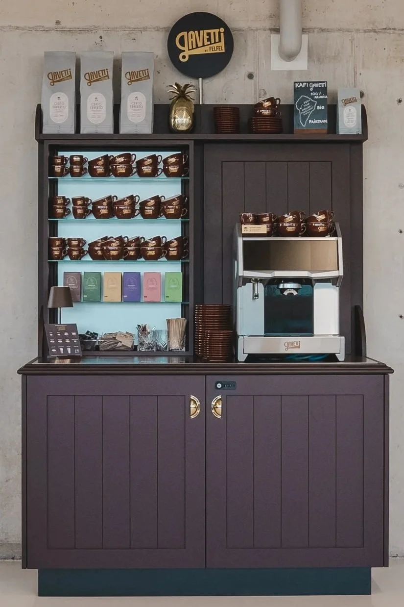 Coffee station with packaged coffee, cups, and a coffee machine, branded 'Gavetti,' set against a concrete wall.