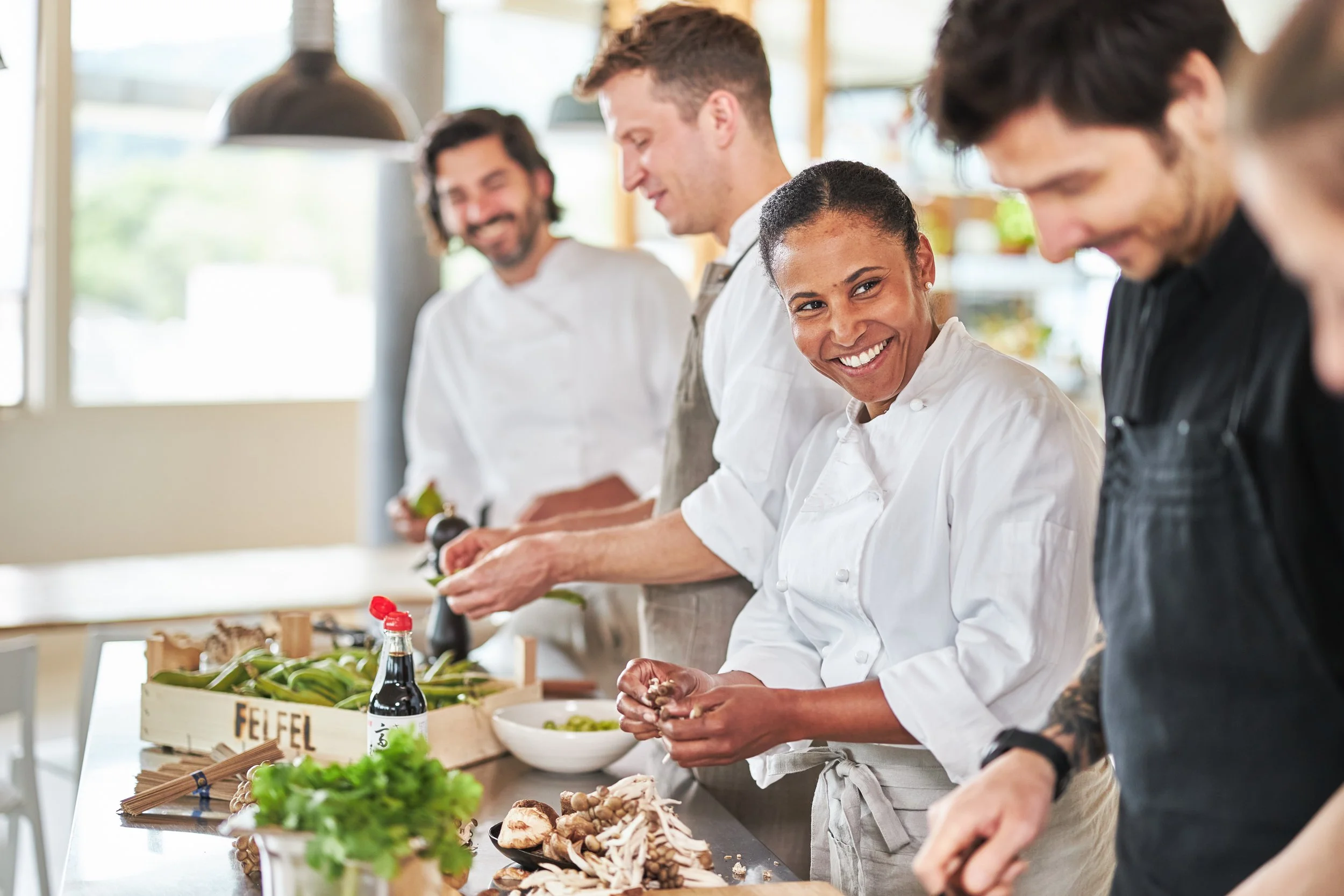 Group of people in chef uniforms preparing food in a kitchen. They are chopping vegetables and smiling.