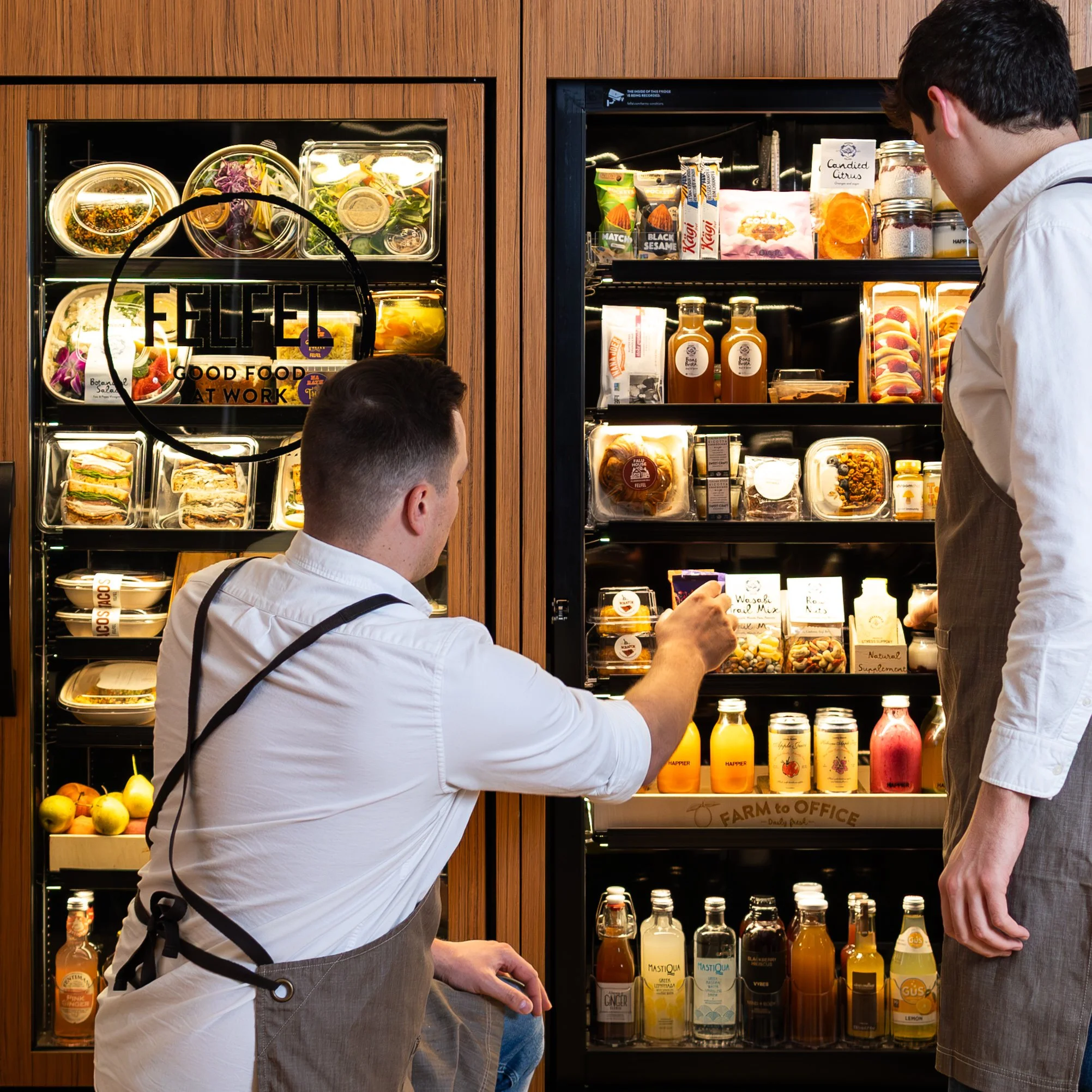 Two women standing in front of a refrigerated vending machine filled with various packaged snacks and beverages in a modern store or café.