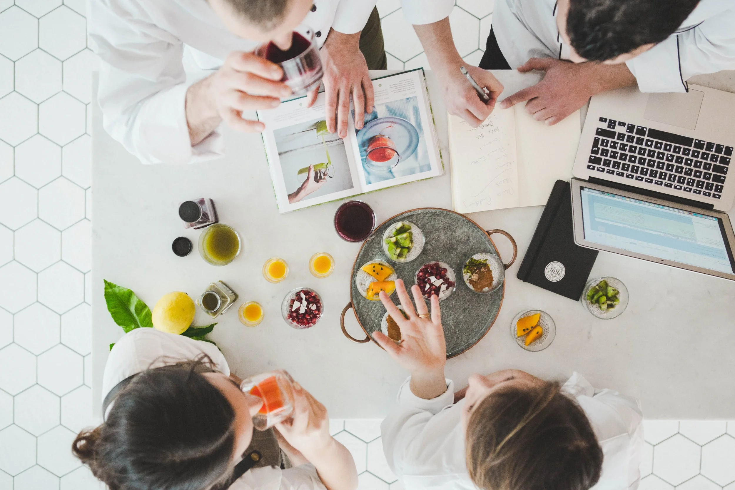 A top-down view of a group of people tasting and discussing various drinks and desserts at a white table, with laptops, notebooks, and colorful food and drink samples.