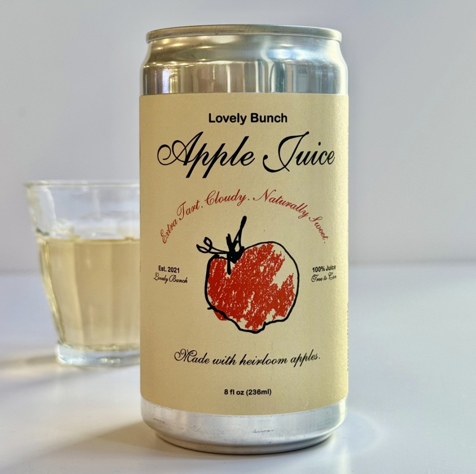 A can of apple juice with a beige label featuring text and a red apple illustration. Next to the can is a glass of light-colored apple juice on a white surface.