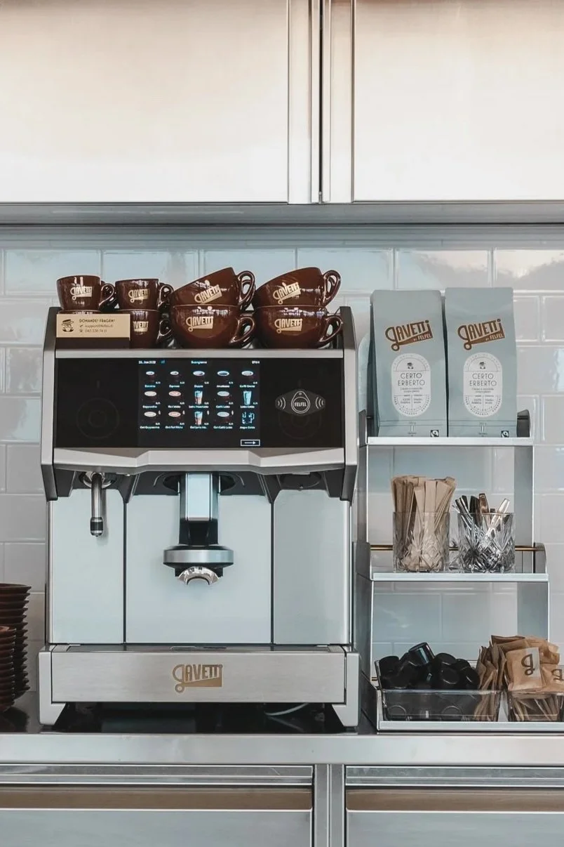 A commercial coffee machine with multiple brown coffee cups on top, branded with Lavazza, and shelves holding coffee supplies like filters, packets, cups, and stirrers.