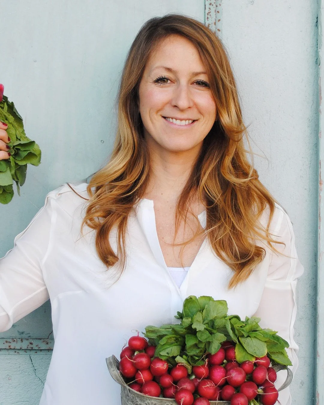 A woman with long, blond hair smiling and holding a basket of fresh radishes with green leaves, standing against a light-colored wall.