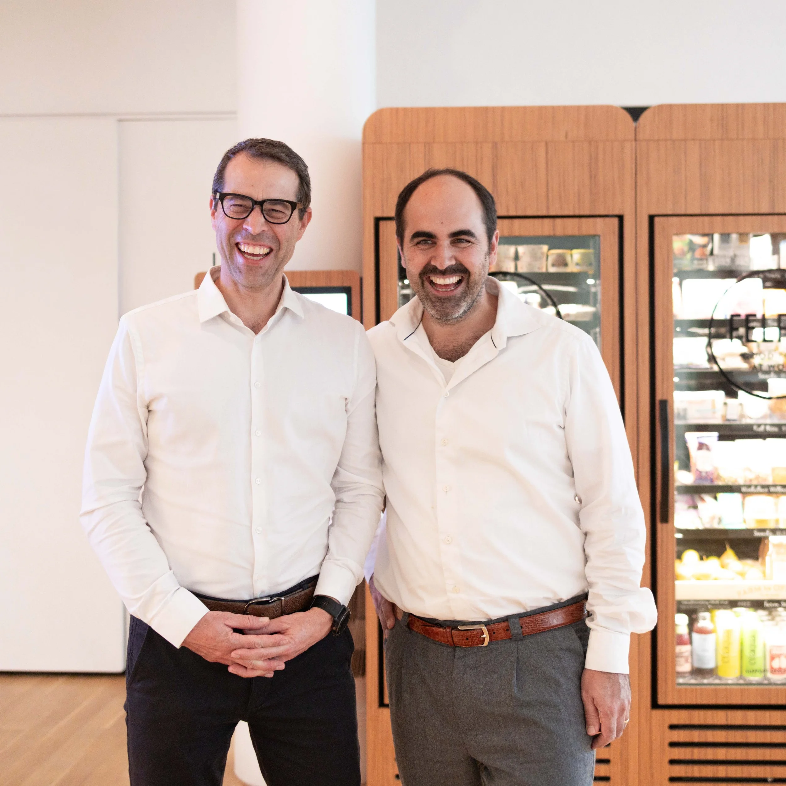 Two men in white shirts smiling and standing together in front of wooden display cases filled with snack items.