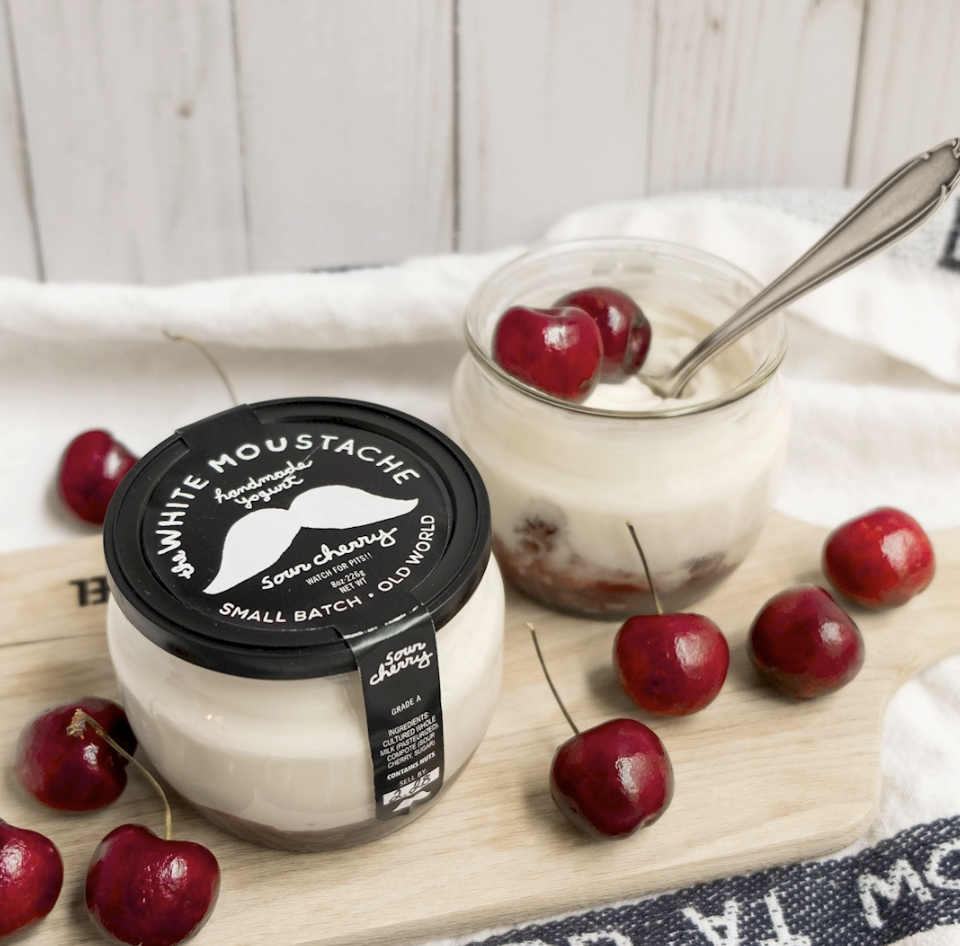 A jar of sour cherry white mustard and a glass jar of sour cherry yogurt with cherries on top and around it, all placed on a wooden cutting board.
