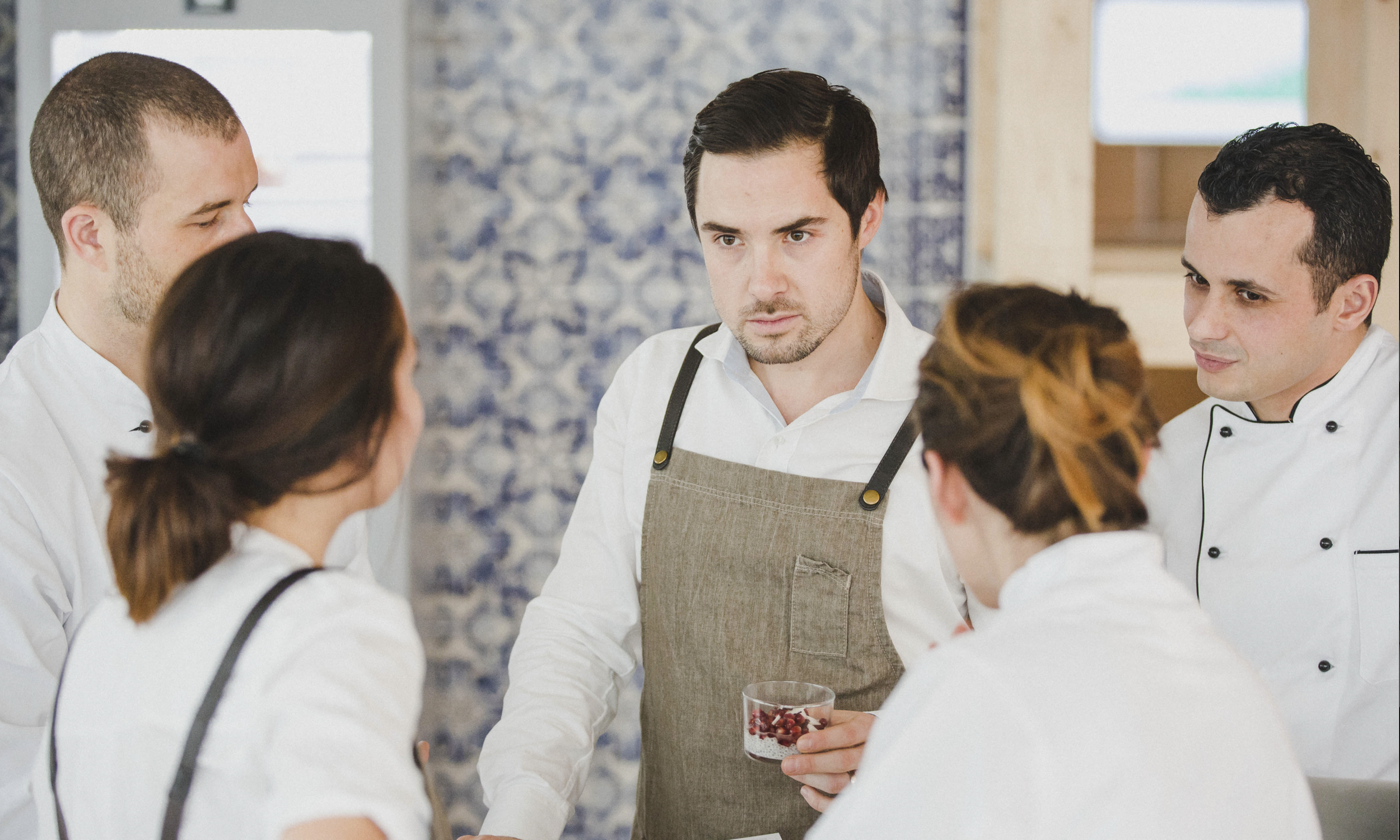 Group of professional chefs in conversation in a modern kitchen, one holding a bowl of red berries.