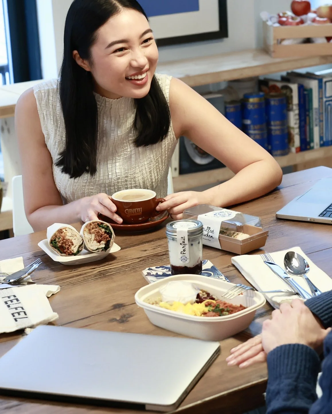 Catering NYC: A woman smiling while holding a coffee cup at a dining table with various food items and a laptop.