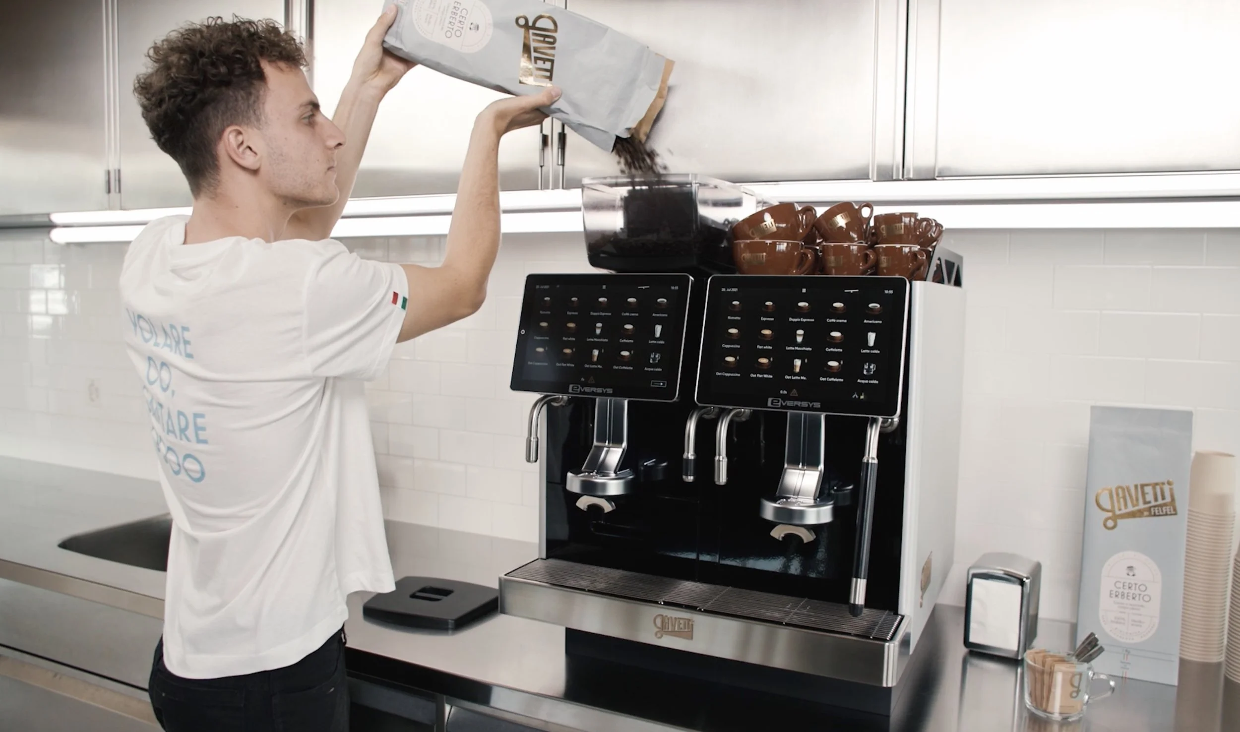 A young man with curly hair is pouring coffee grounds into a coffee machine in a modern kitchen or coffee shop. The coffee machine has digital touchscreens and multiple spouts, with cups placed on top. There are paper cups, a box, and a small container on the counter nearby.