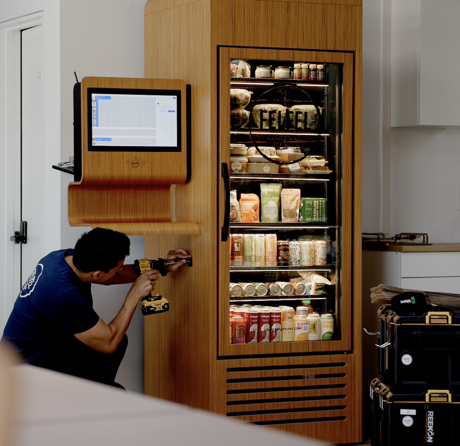 A man in a blue shirt is using a cordless drill to install or repair a wooden-paneled refrigerator filled with various food and beverage products, in a modern kitchen with white cabinets.